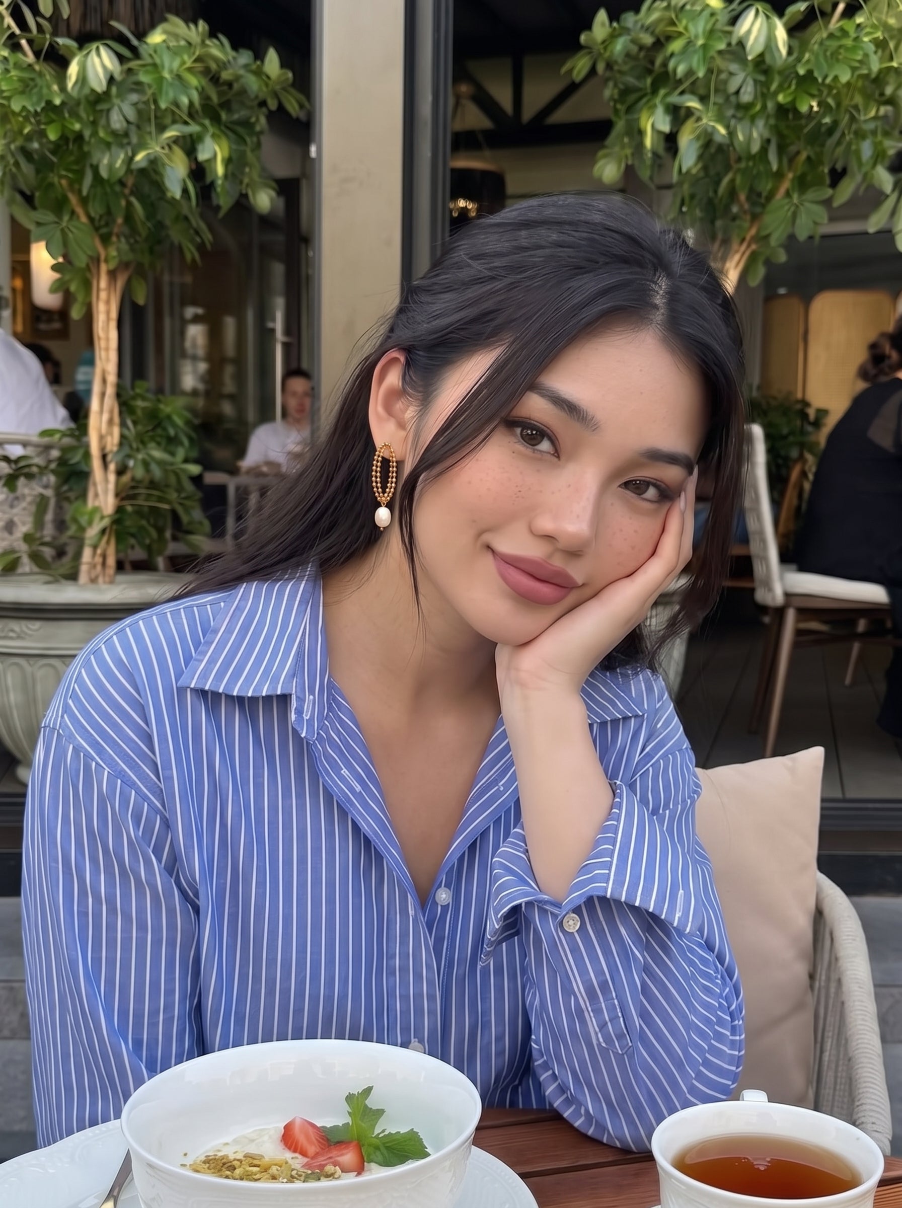 A woman wearing Aperture Freshwater Pearl Drop Earrings while enjoying a meal outdoors. The earrings feature a gold-tone oval bead design with a single white pearl. @lifestyle