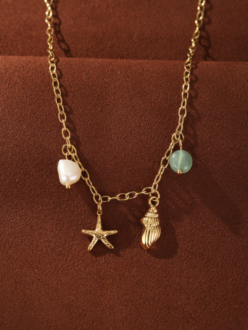 Collier doré avec une chaîne délicate présentant une perle baroque d'eau douce, une perle verte, une breloque étoile de mer dorée, et une breloque coquillage dorée.