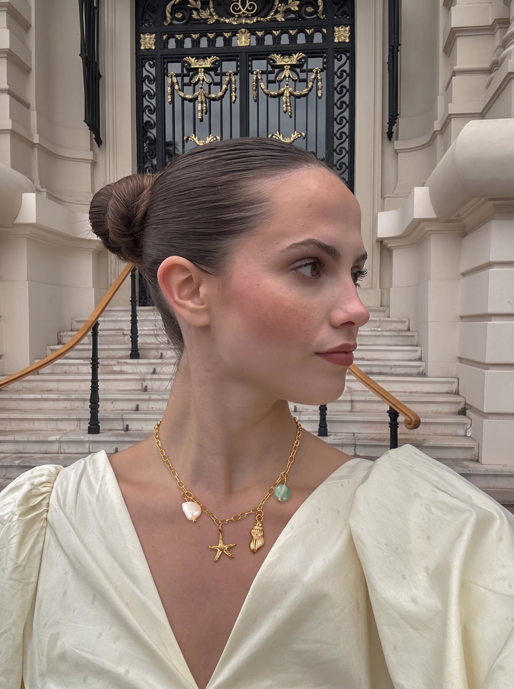 Femme portant le collier Aqua Radiance avec des breloques en perle d'eau douce, coquillage, et étoile de mer, stylisé avec une robe crème à manches bouffantes dans un cadre extérieur avec de grands escaliers et de l'architecture. @lifestyle