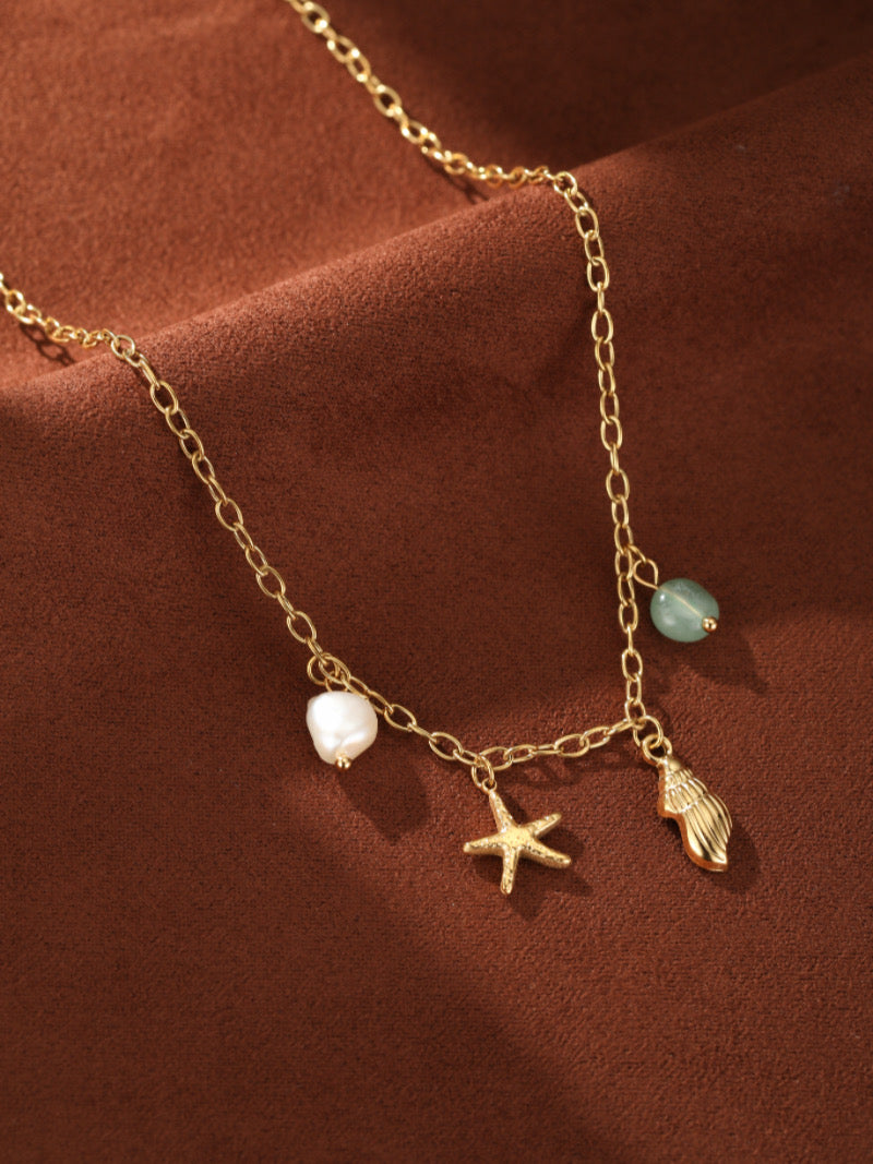 Collier à chaîne dorée présentant une perle baroque d'eau douce, une breloque étoile de mer dorée, une breloque coquillage dorée, et une perle bleu clair.