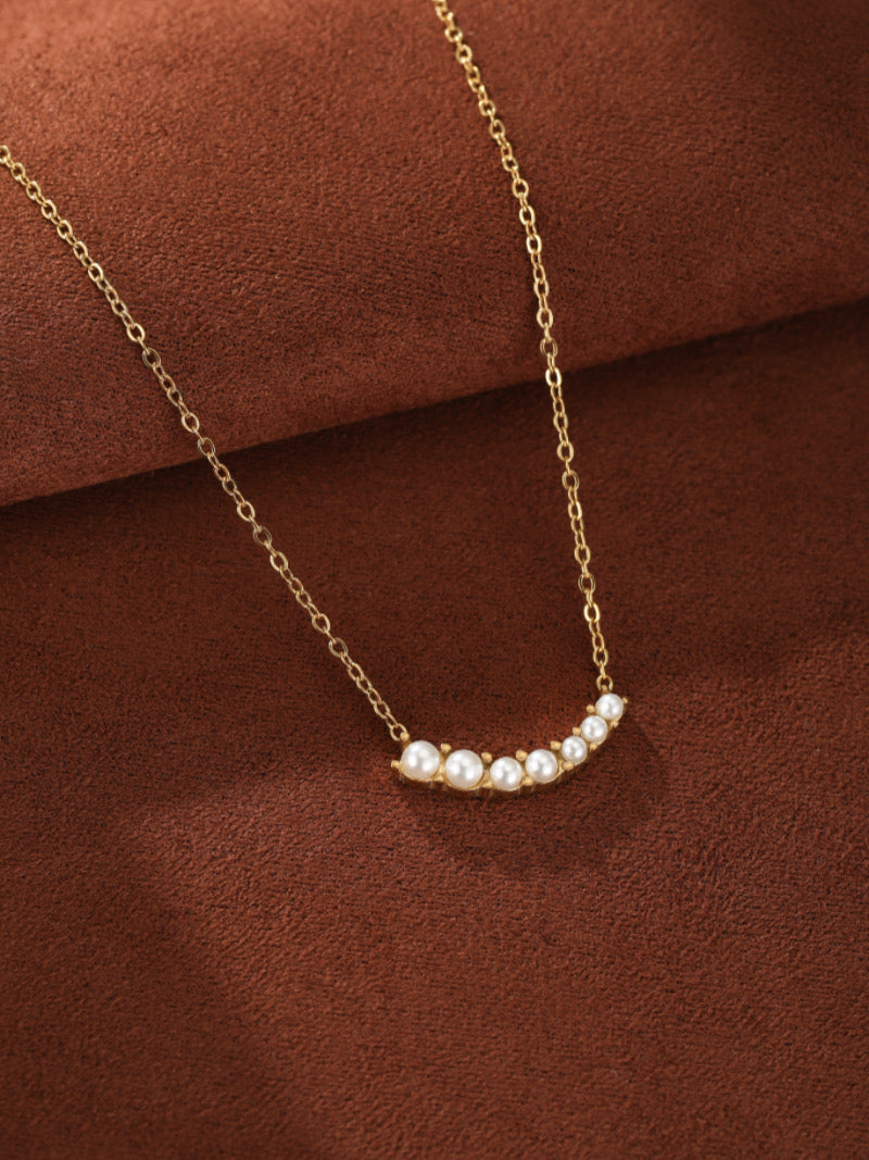 Gold necklace with a curved bar featuring seven small, luminous white freshwater pearls on a brown textured background.