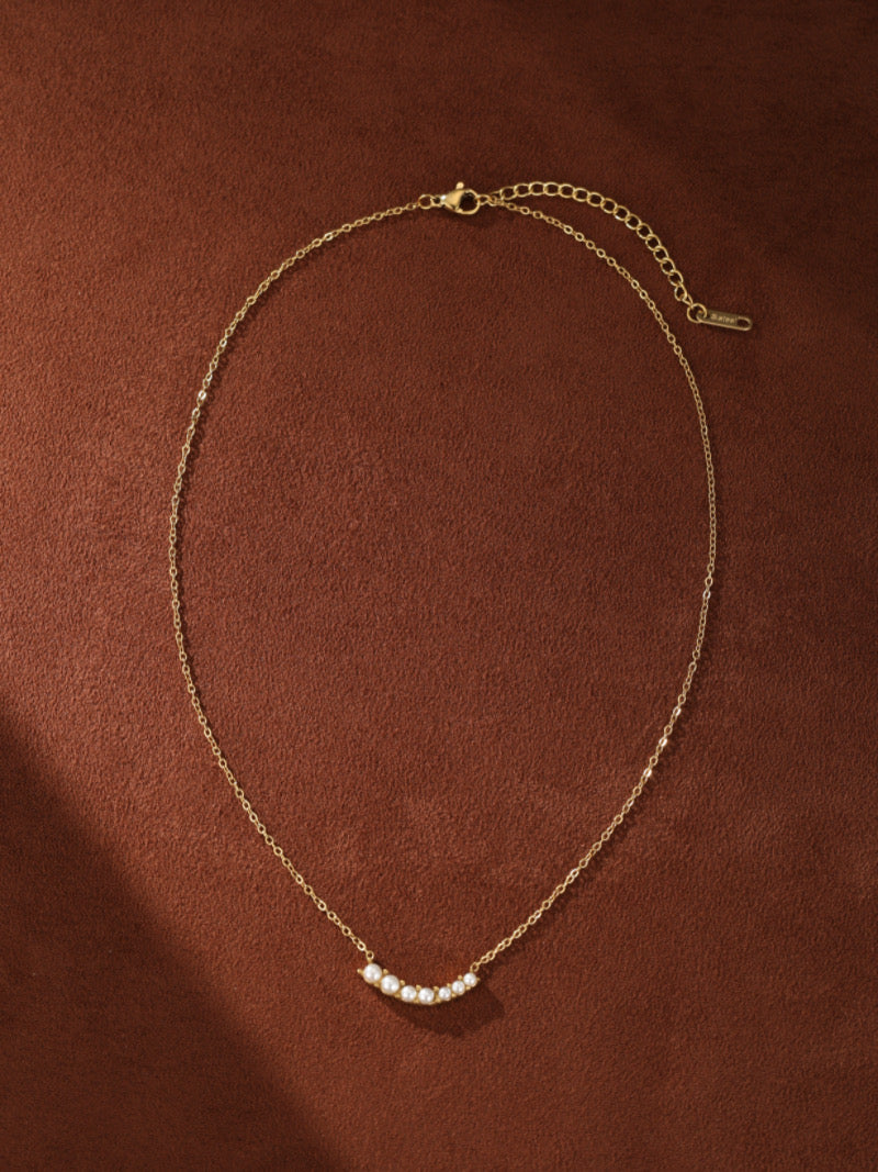 Gold necklace with a curved bar featuring seven small, round, white freshwater pearls on a brown background.