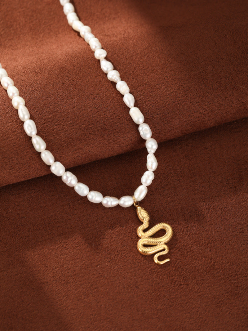 Un collier pendentif serpent ton or avec des perles blanches irrégulières d'eau douce enfilées sur une chaîne fluide, présenté sur fond texturé marron.