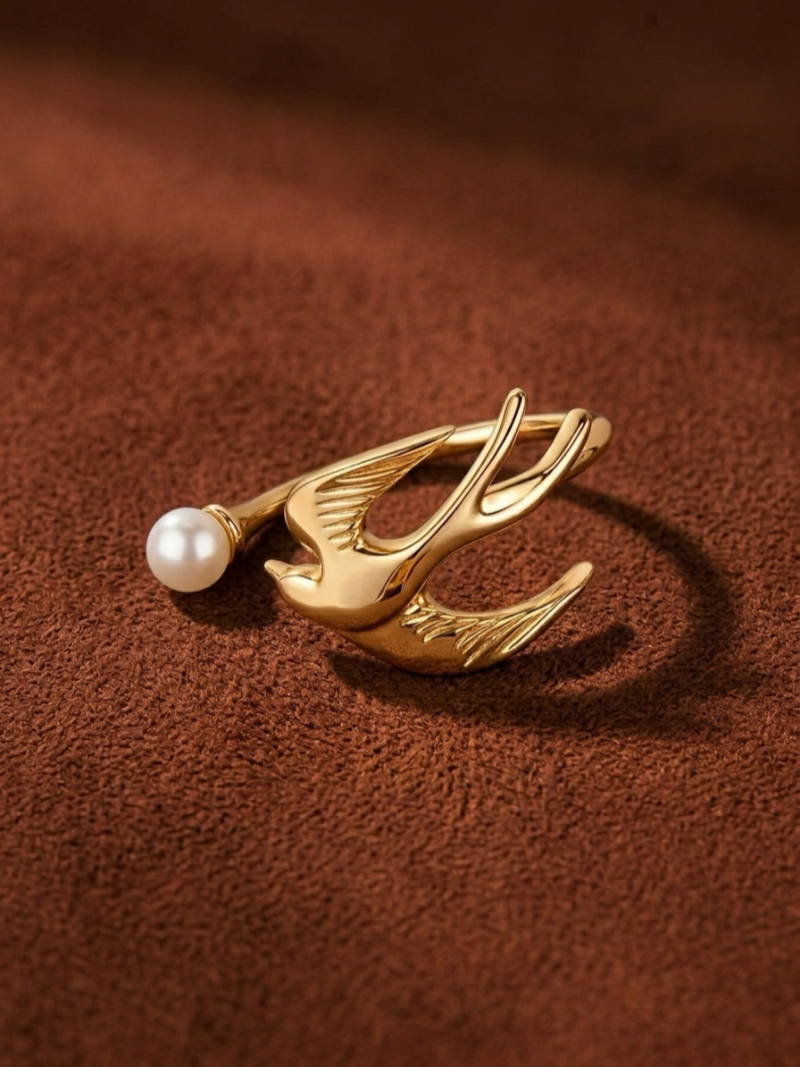 Gold-tone cuff ring with a sculpted bird design and a small freshwater pearl on the left, set against a dark brown textured background. @pearl