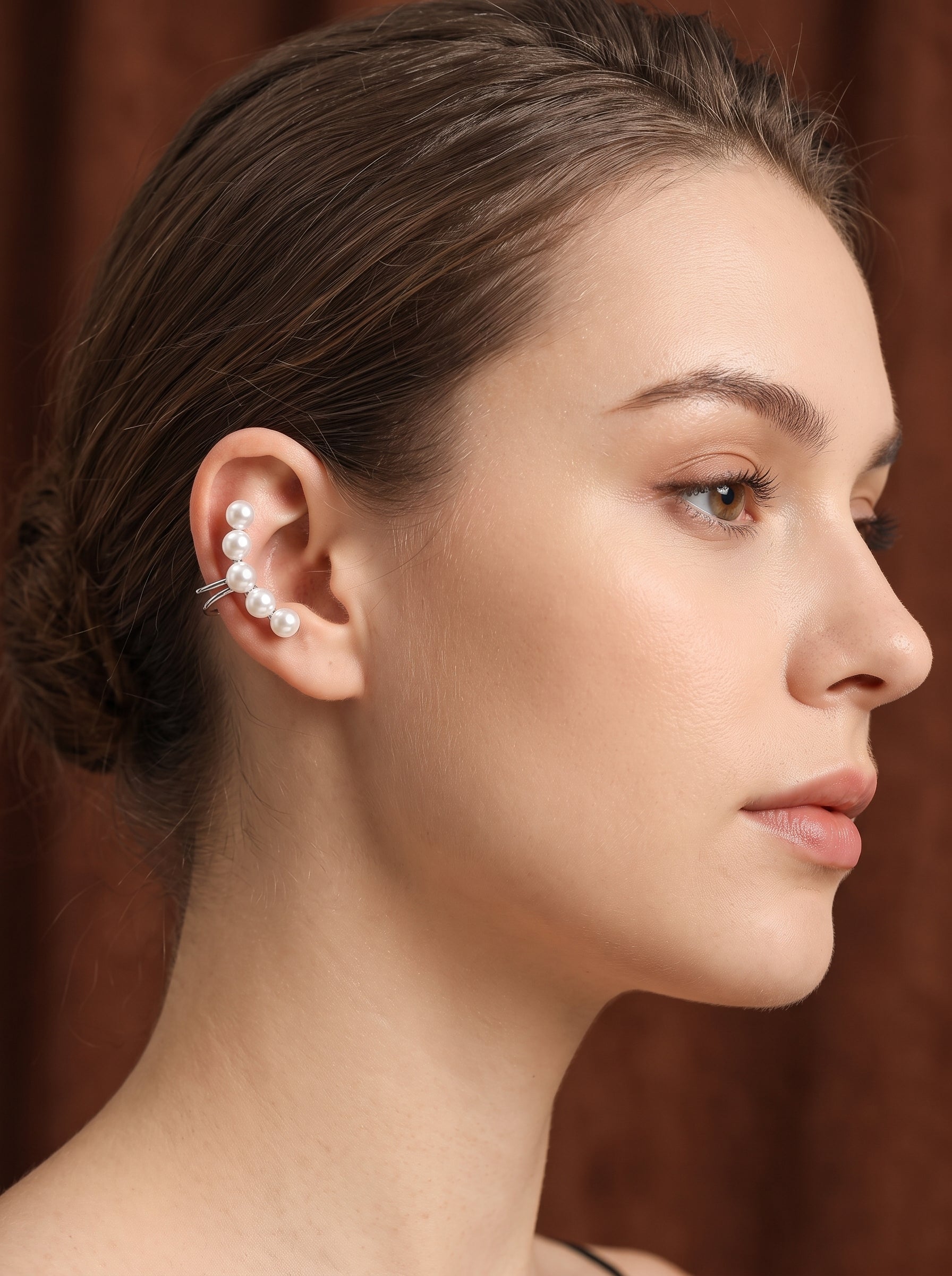 Une mannequin porte les boucles d'oreilles créoles Ascent en perle d'eau douce, présentant une rangée courbée de cinq lustrées perles blanches rondes de 5 mm remontant le lobe de l'oreille sur fond marron. @model @lifestyle