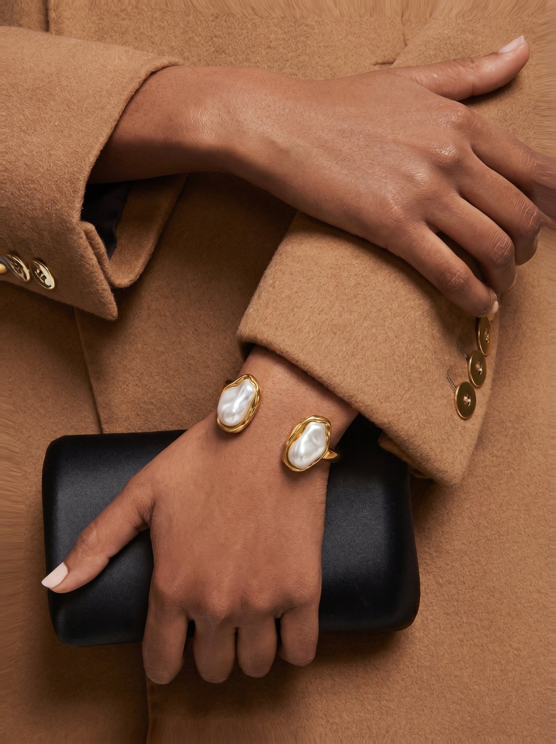 Close-up of a woman's wrist adorned with the Aura Freshwater Pearl Cuff Bracelet, showcasing two irregular white Baroque pearls set in a gold-toned open cuff, held against a black clutch and a tan coat. @model