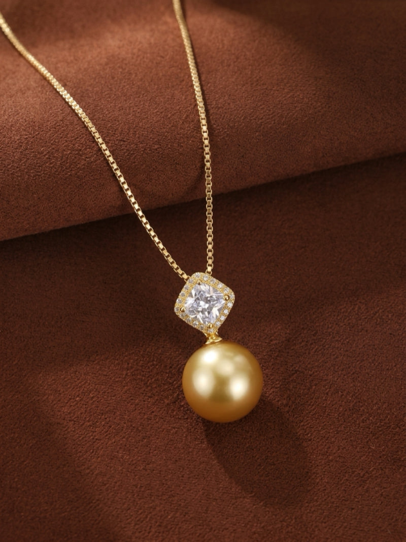 Aurelia single pendant necklace featuring a large, round 15mm South Sea Golden Pearl suspended from a diamond-accented square bail on a delicate gold chain, displayed on a brown textured background.