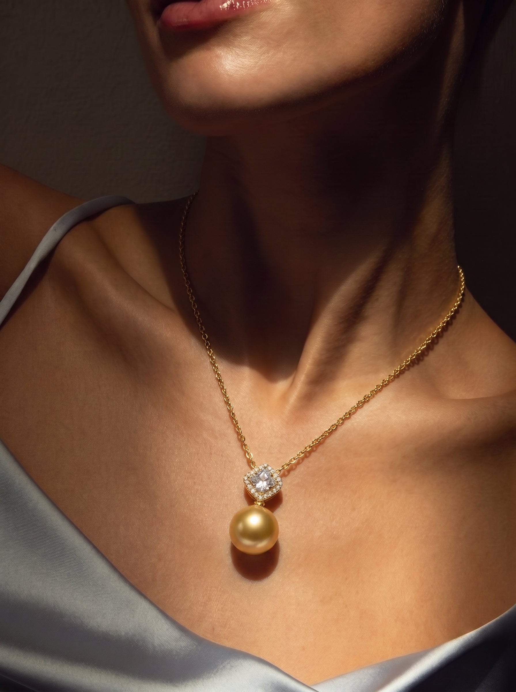 A close-up shot of a woman wearing the Aurelia South Sea Pearl Square Single Pendant Necklace, featuring a lustrous 15mm golden pearl suspended from a square bail adorned with accent stones, on a delicate gold chain. @model