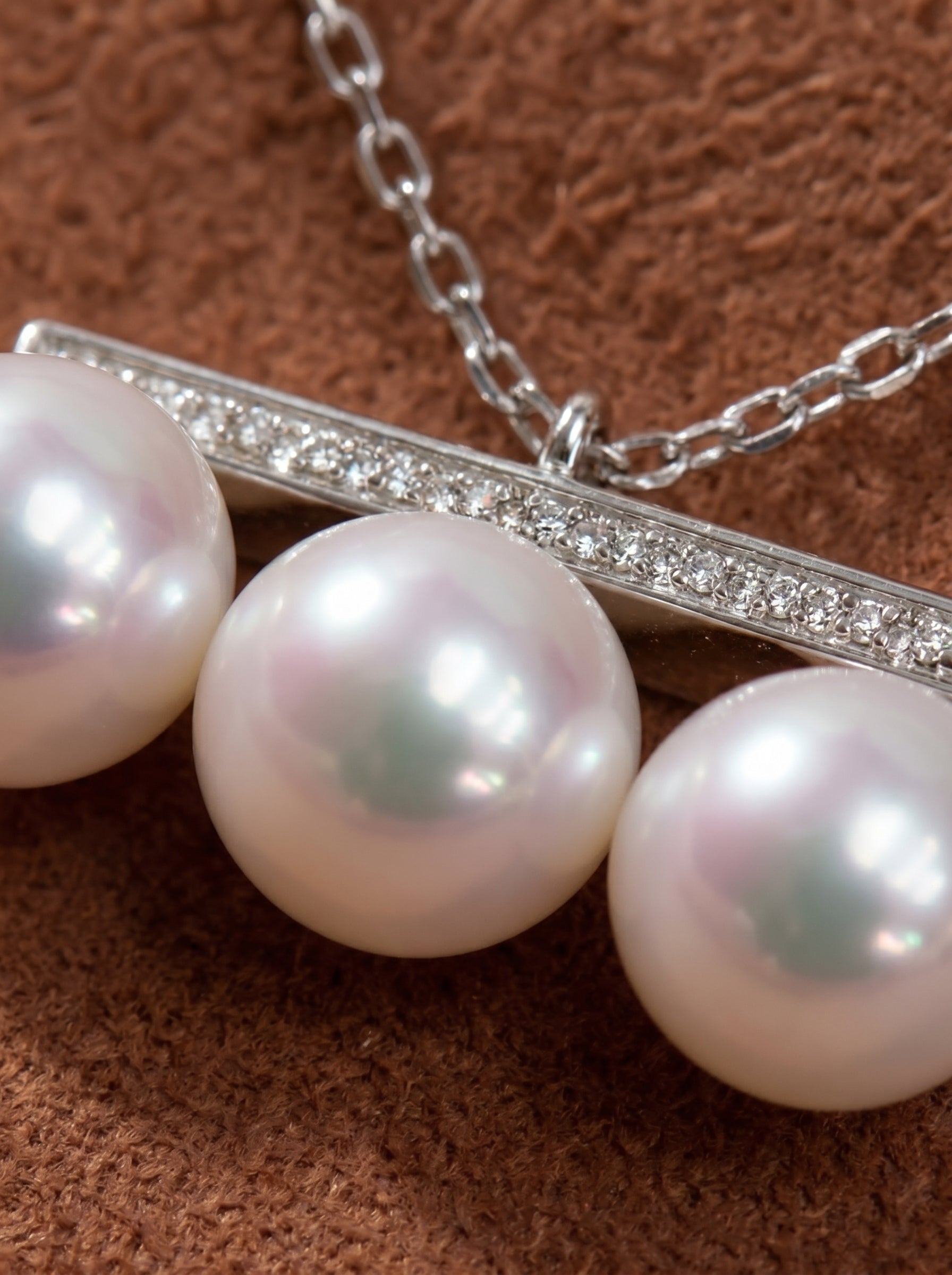 Close-up of a white gold chain necklace featuring a pave diamond bar pendant that suspends three luminous 7mm Akoya white pearls with subtle pink and green iridescence. @pearl