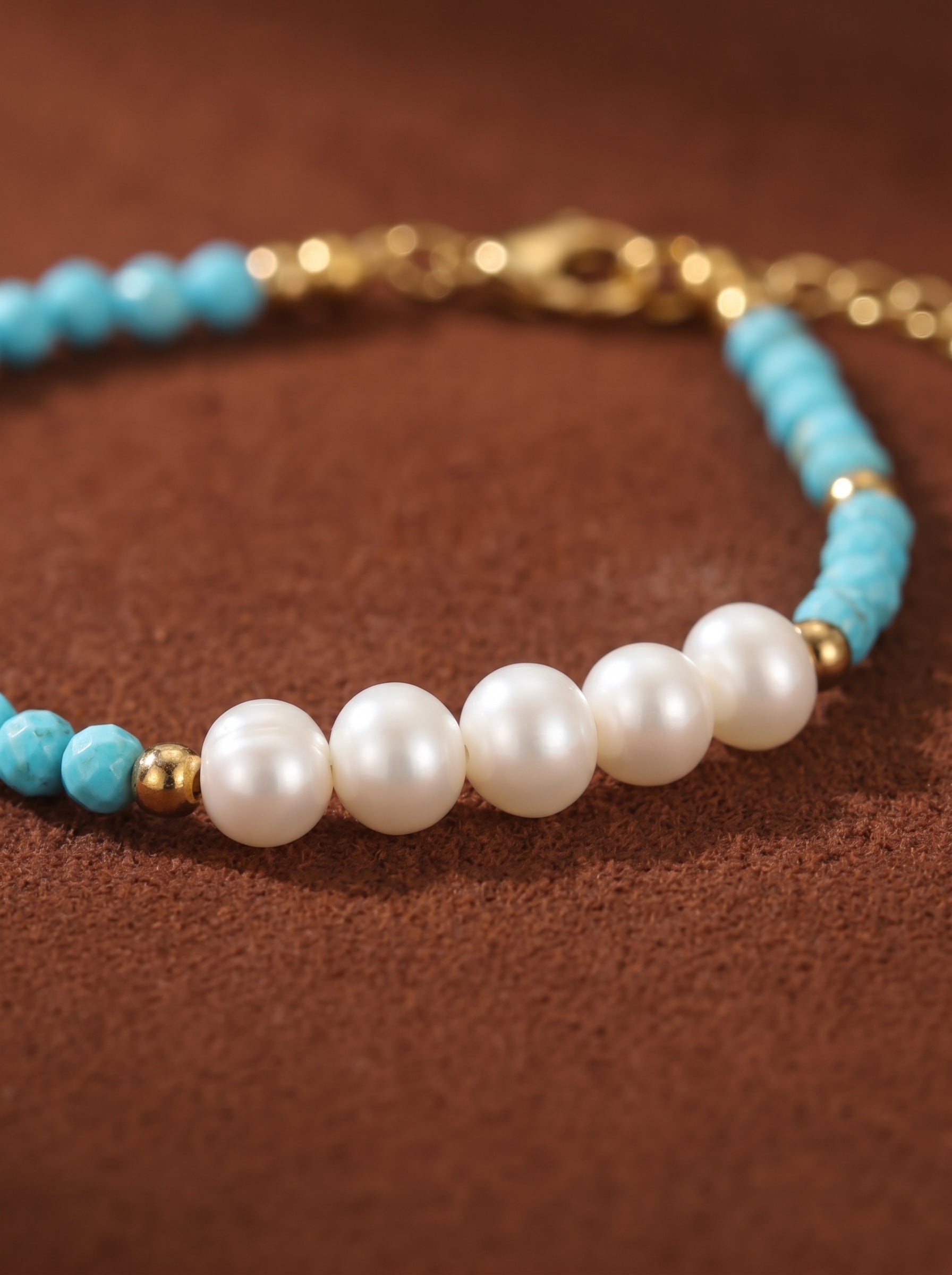Close-up of a bracelet with white freshwater pearls, small turquoise beads, and gold spacers on a brown background. @pearl