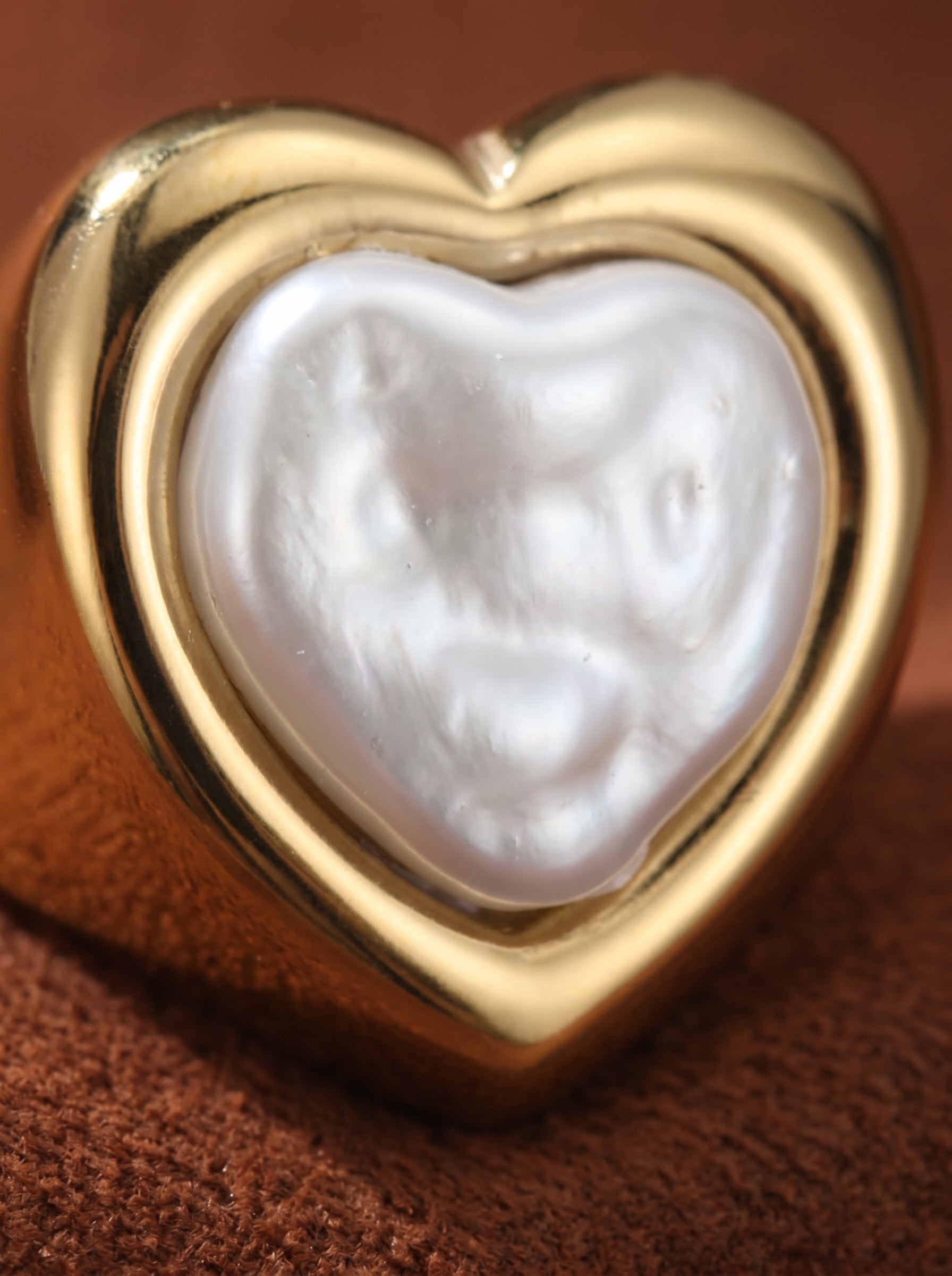 Close-up of a bold gold-tone cuff ring featuring a heart-shaped bezel set with an irregular white freshwater pearl. The ring has a polished finish and rests on a textured brown surface. Freshwater Pearl Heart Cuff Rings@pearl