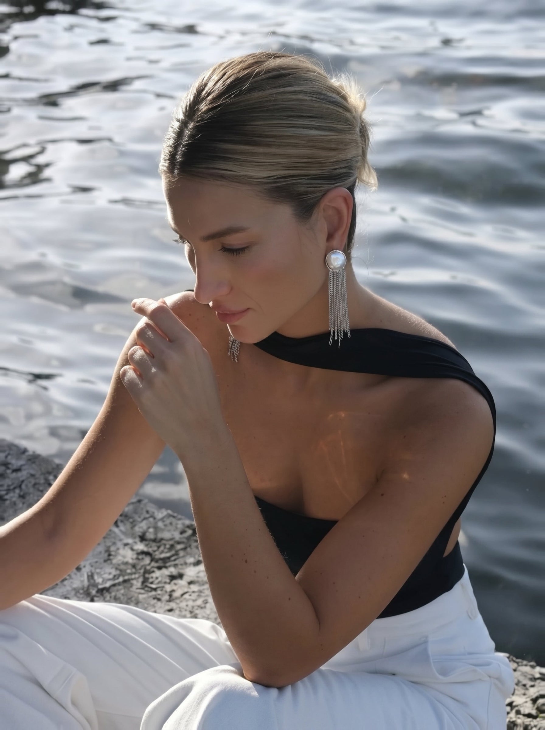 A woman sits by the water wearing the Cascade Freshwater Pearl Fringe Dangle Earrings, featuring a large baroque pearl and silver-toned chain fringe. @lifestyle