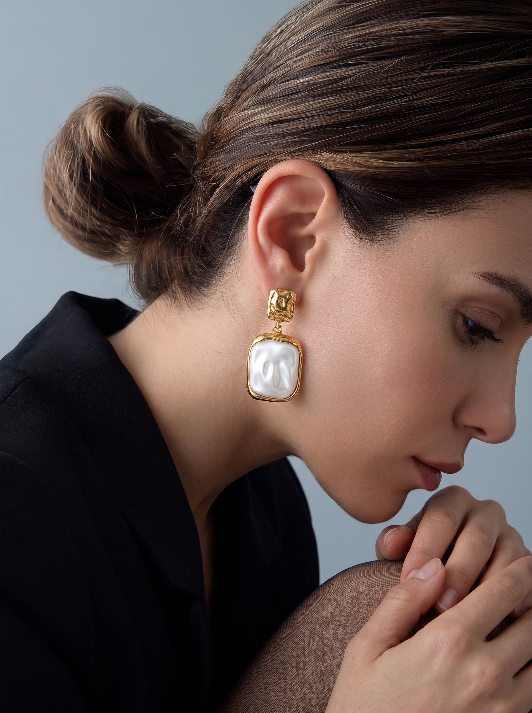 Gros plan d'une femme portant les boucles d'oreilles pendantes relief Cast en perle d'eau douce, présentant une grande et irrégulière perle baroque blanche dans une monture carrée texturée dorée, suspendue à une puce carrée martelée. @model