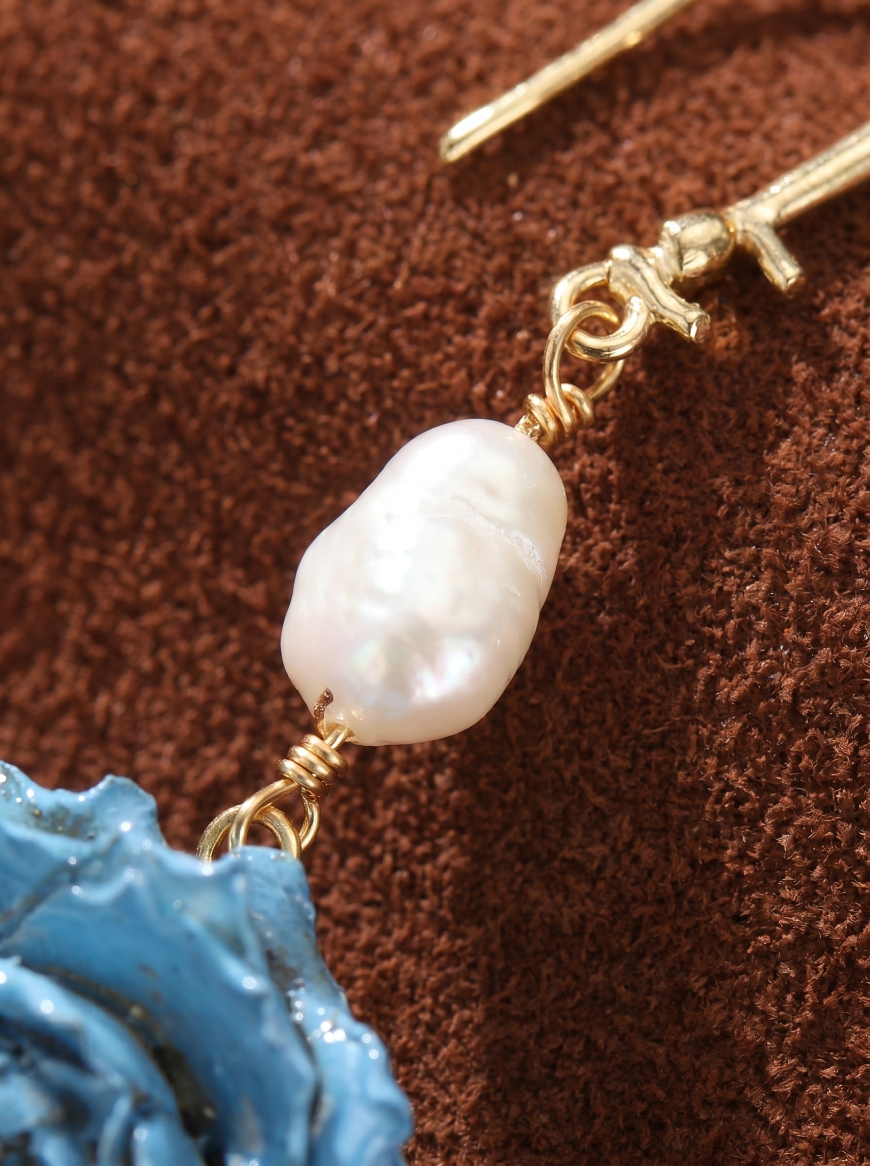 Close-up of Celeste Reverie dangle earrings featuring a textured blue flower motif and a baroque white freshwater pearl hanging from a gold hook.@pearl