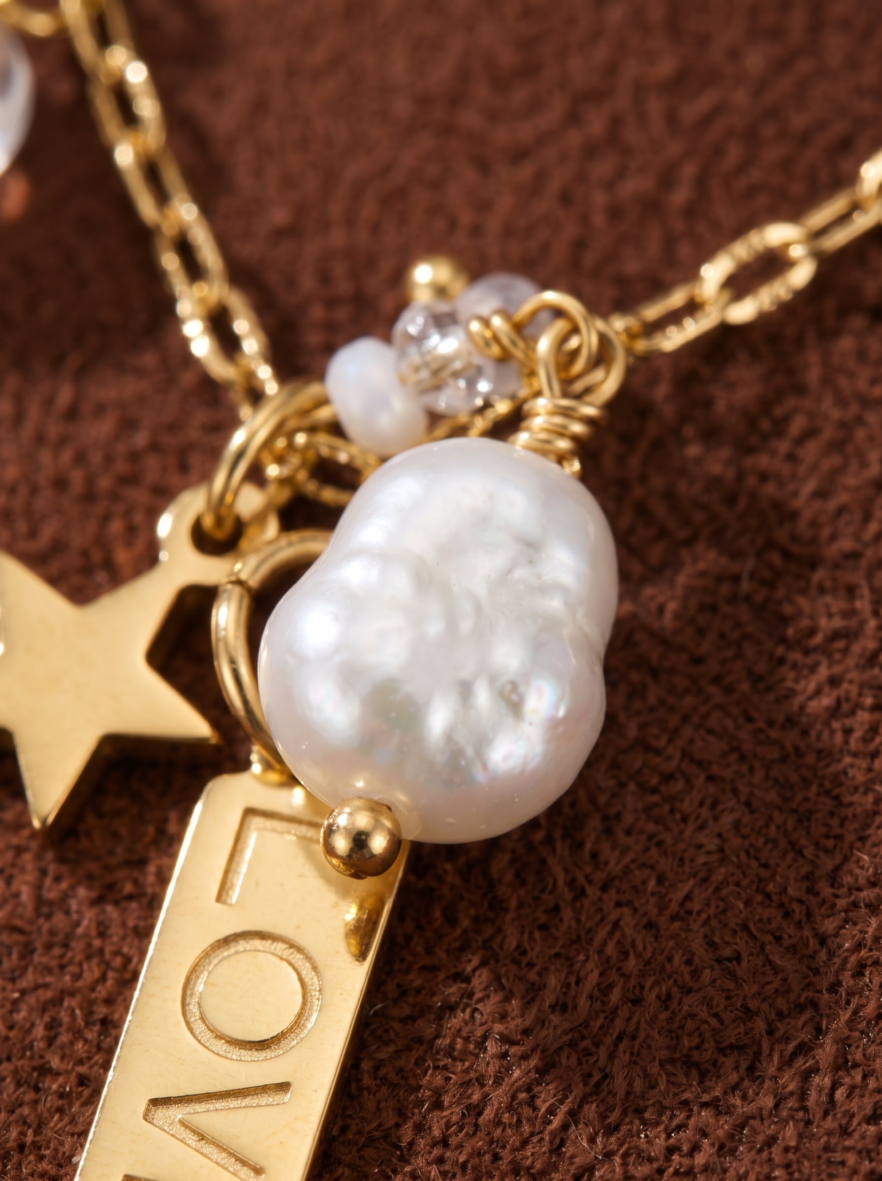 Close-up of a gold-tone necklace featuring a large, irregular white freshwater baroque pearl, a gold star charm, and a rectangular tag engraved with the word 'LOVE'. The necklace chain is delicate and textured. @pearl
