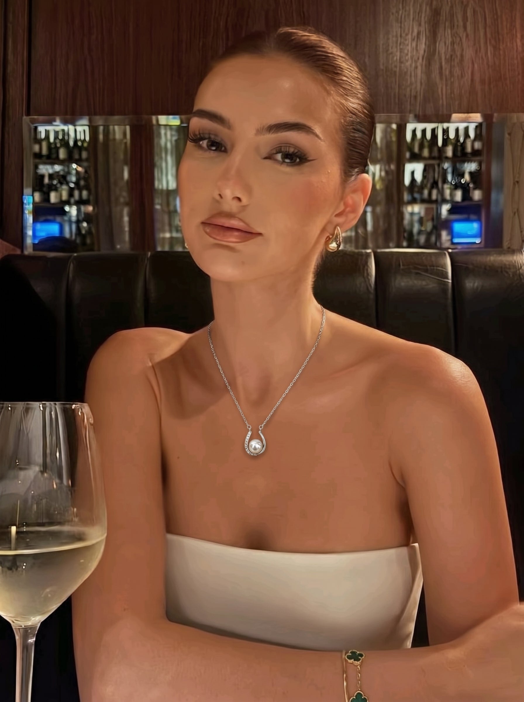 A woman with styled hair and makeup wears a silver-toned chain necklace featuring a horseshoe pendant with a luminous Akoya pearl at its center, and matching hoop earrings, while seated at a table with a glass of white wine. @model #model #lifestyle