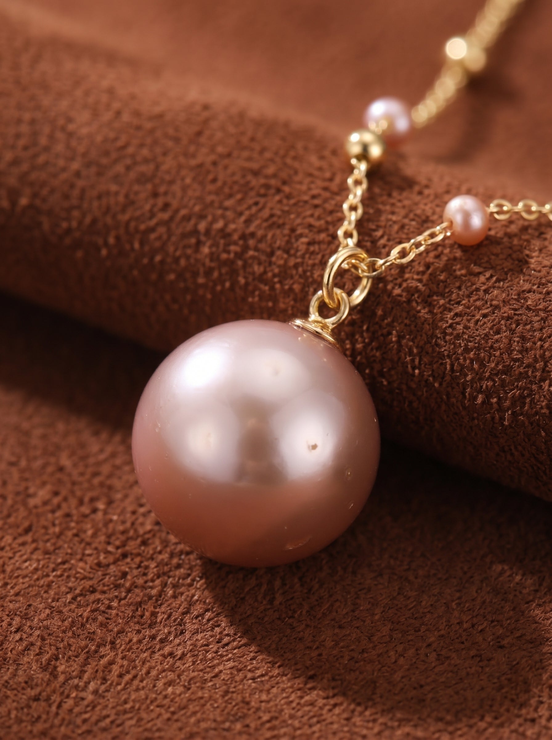 Close-up of a Chiffon Freshwater Pearl Floating Pendant Necklace featuring a large, lustrous pink pearl as the centerpiece, with smaller pink pearls and gold beads scattered along the delicate gold-tone chain, set against a textured brown background.@pearl