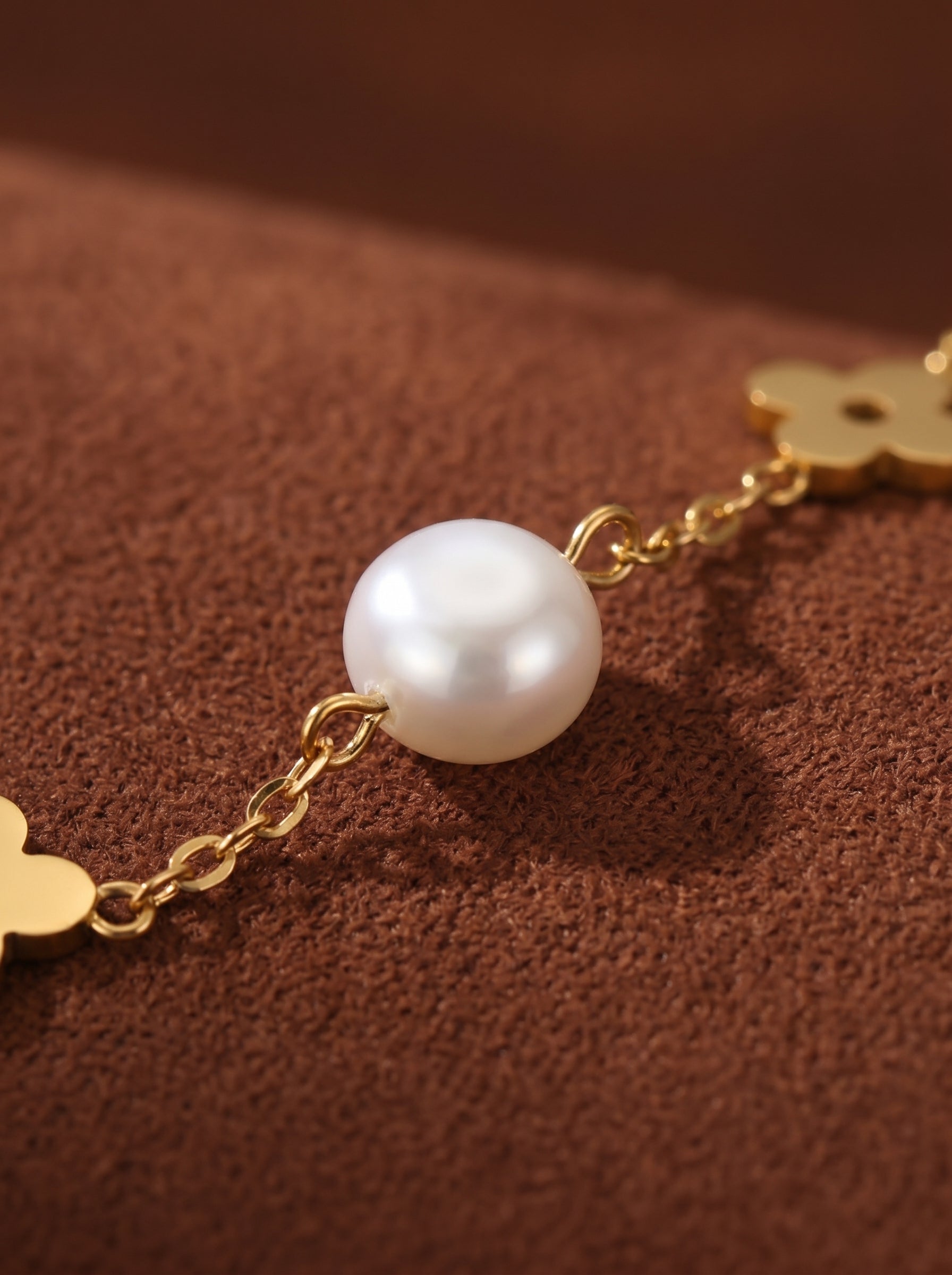 A close-up of a gold chain bracelet featuring a lustrous white freshwater pearl and small gold clover charms on a brown textured background. @pearl