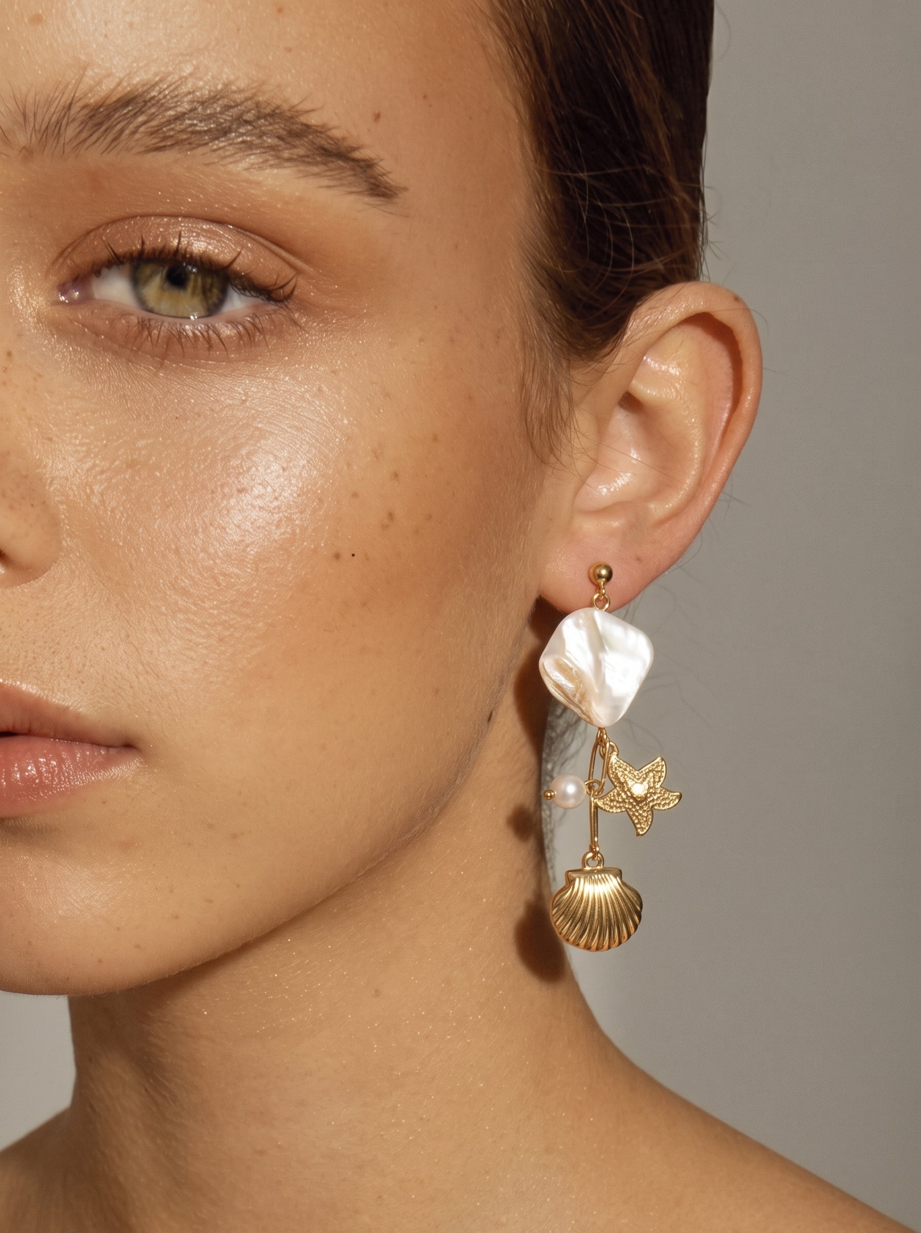 Gros plan d'une mannequin portant les boucles d'oreilles Coastal Allure présentant une grande et irrégulière pièce en nacre, une petite perle d'eau douce, une breloque étoile de mer dorée, et une breloque coquillage dorée pendant d'une tige dorée. @model