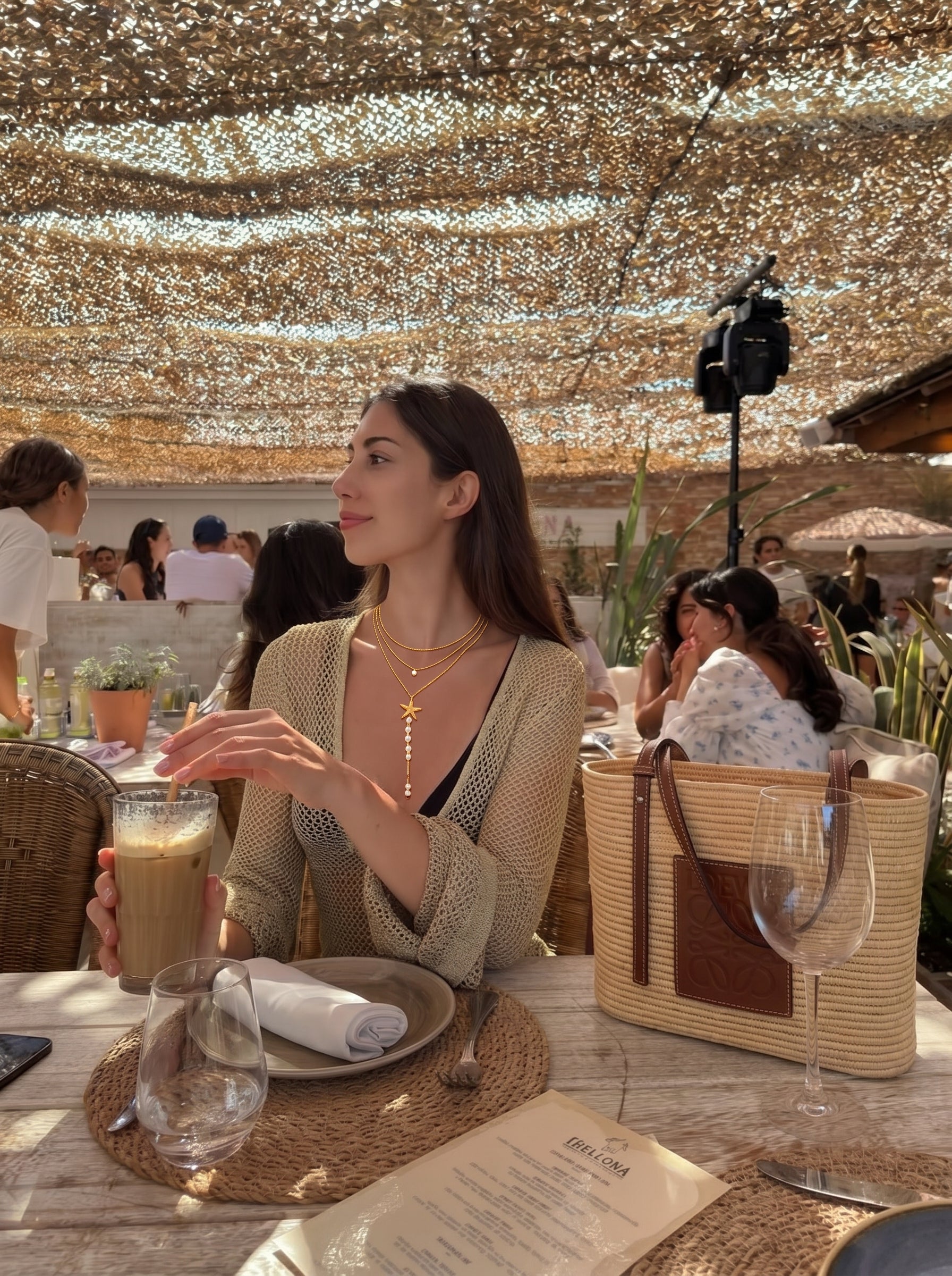 Une femme dans un café en extérieur porte le collier superposé long étoile Coastal Gleam en perle d'eau douce, présentant de délicates chaînes dorées, une breloque étoile de mer, et de lumineuses perles graines blanches d'eau douce. @lifestyle