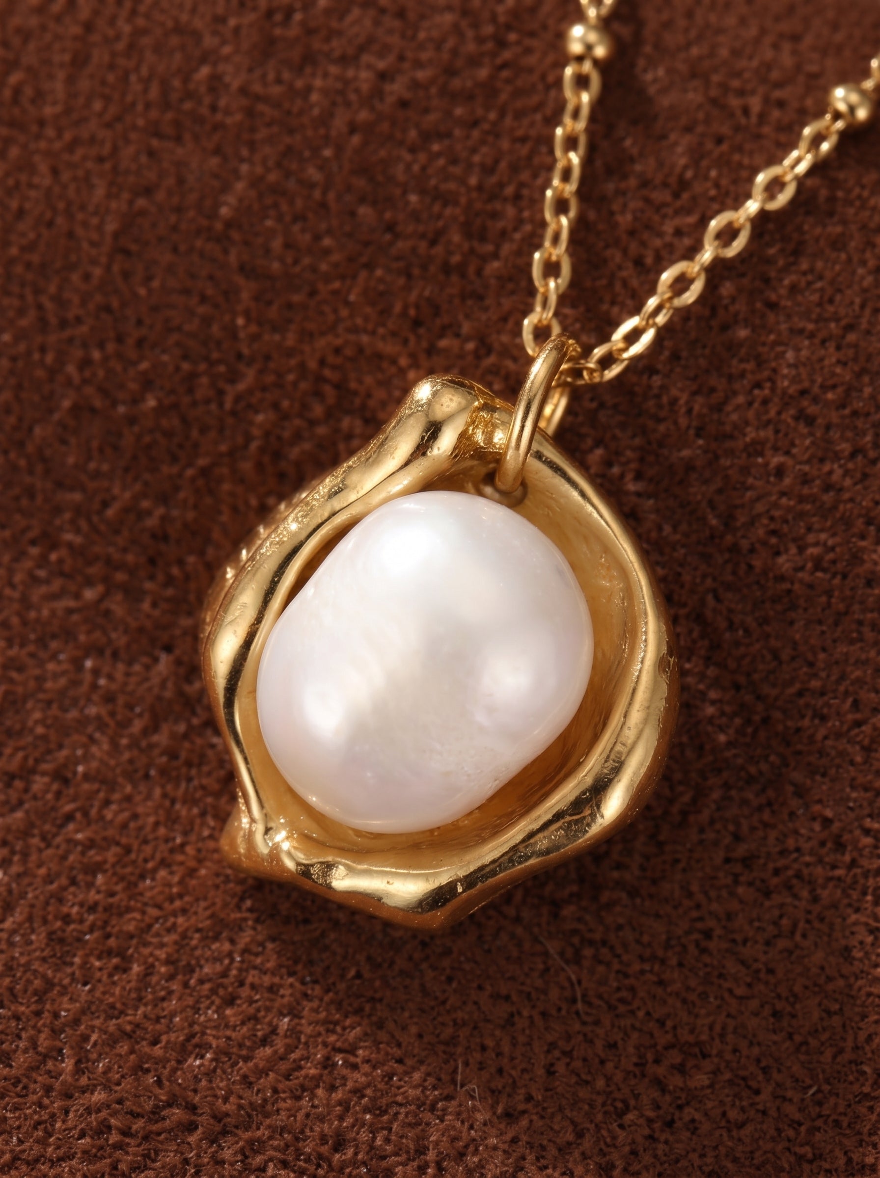 Gros plan du pendentif du collier Cocoon présentant une lumineuse perle blanche d'eau douce nichée dans un vase sculptural ton or, sur une délicate chaîne perlée ton or. @pearl