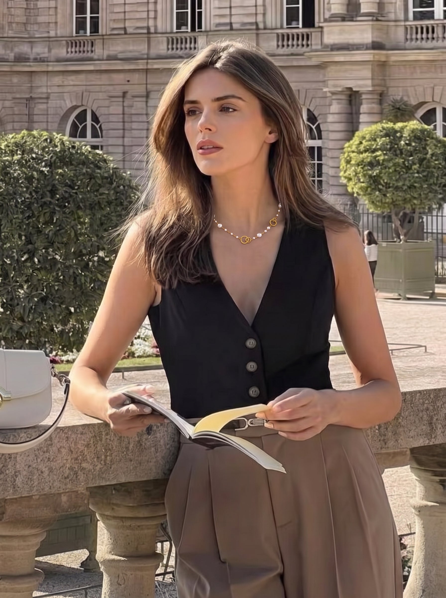 A woman wearing a black vest and brown pants, adorned with the Constellation Imitation Pearl Ring Long Floating Necklace, holds an open book while leaning on a stone railing outdoors near a building and trees. @lifestyle