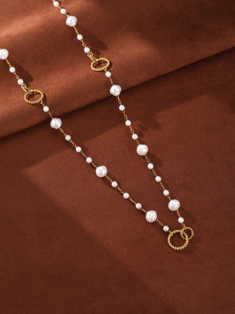 A long, gold-tone chain necklace with varying sizes of white imitation baroque pearls and textured gold rings, laid on a brown fabric surface.