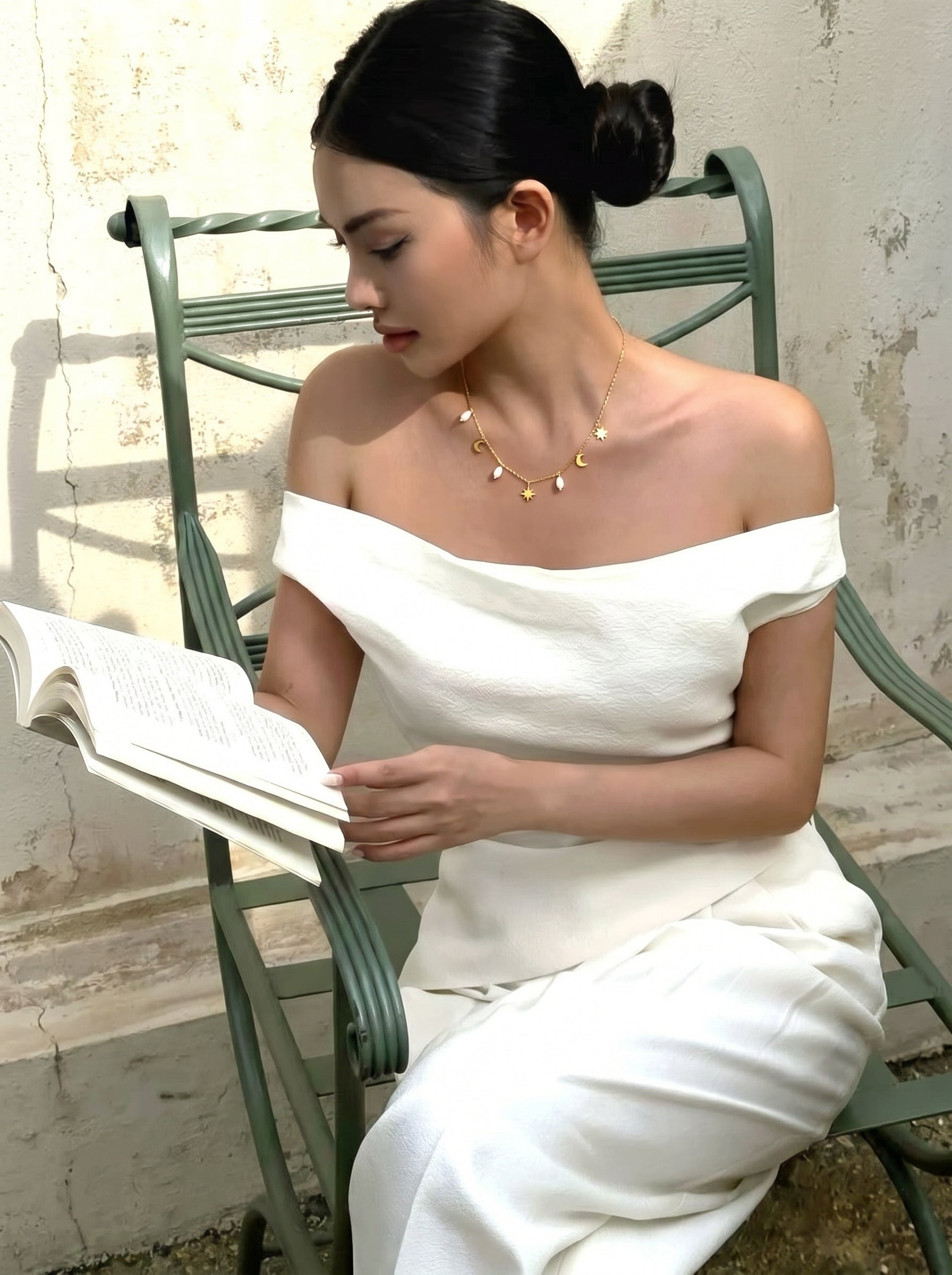 A woman wearing a white off-the-shoulder top and pants sits on a green chair outdoors, reading a book and wearing the Cosmic Whisper necklace with pearl, moon, and star charms. @lifestyle #lifestyle