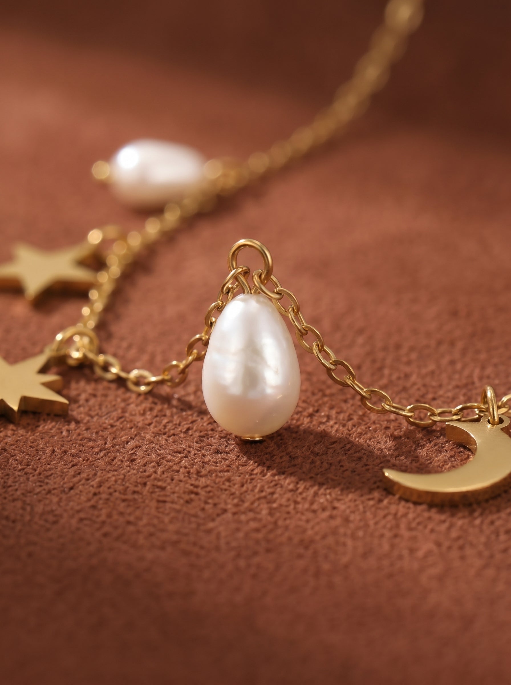 Close-up of a gold chain necklace with a teardrop freshwater pearl and celestial charms including a crescent moon and stars on a brown textured background. @pearl