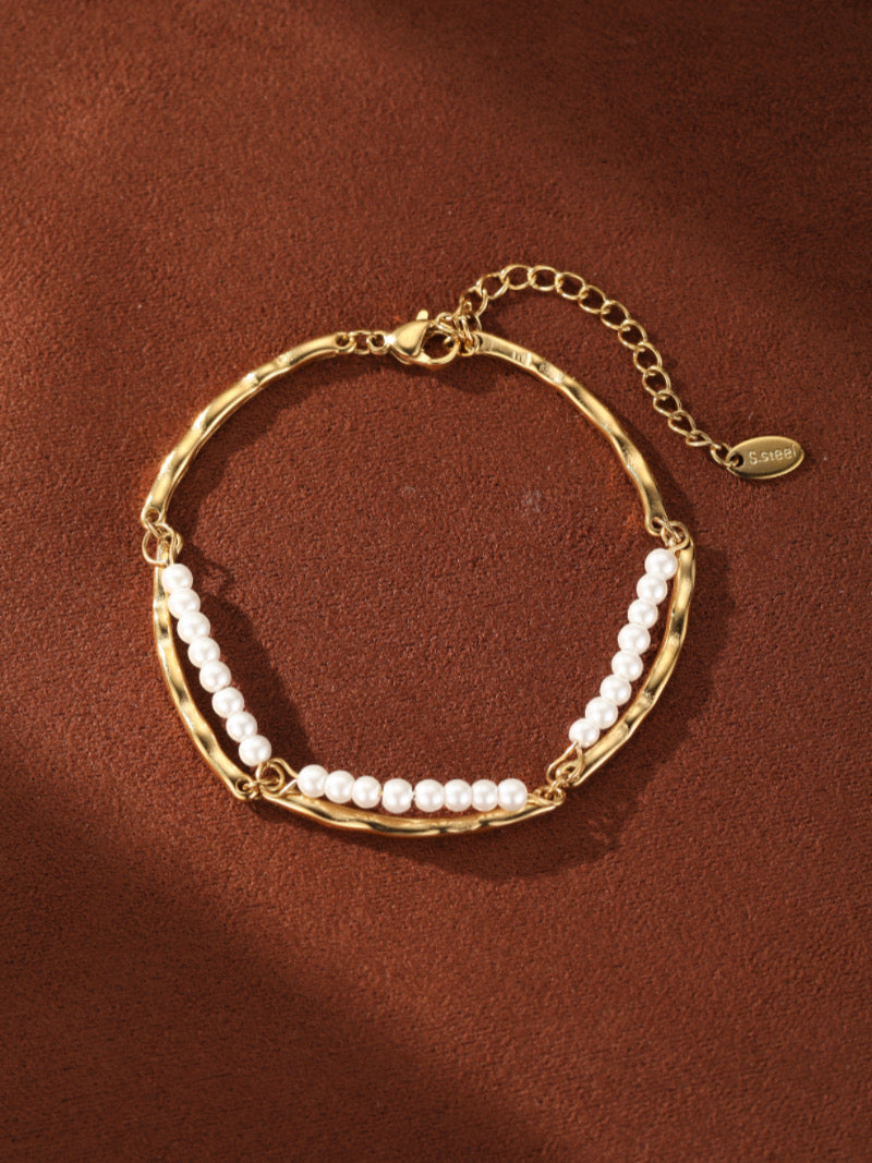 Gold-tone curved chain bracelet with two rows of small white freshwater pearls, on a brown textured background.