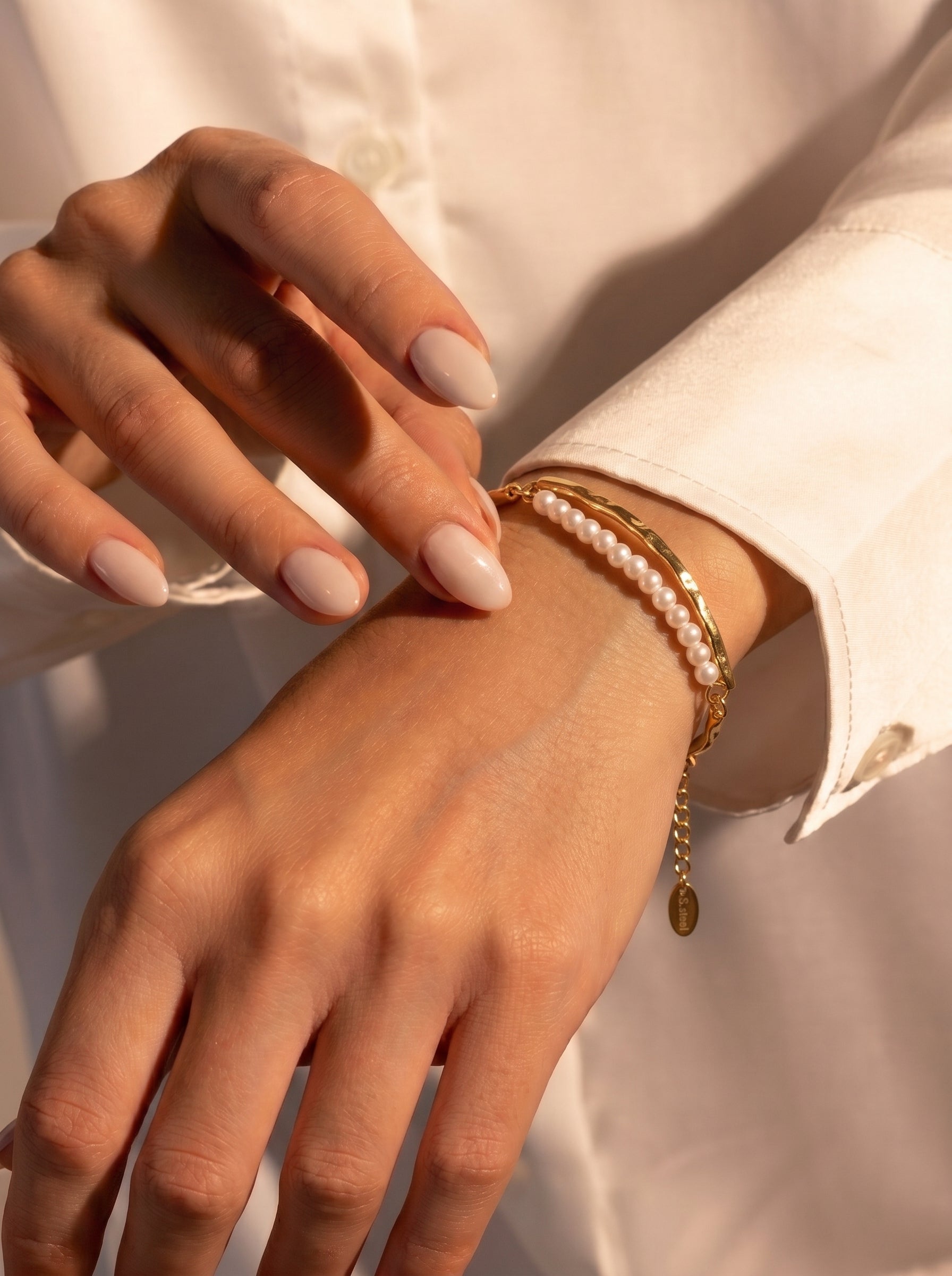 A close-up shot of a woman's wrist wearing the Covelet Freshwater Pearl Curved Chain Bracelet. The bracelet features a gold-tone curved band with two delicate rows of small, white freshwater pearls and a fine gold chain. The woman has neatly manicured nails in a neutral shade. @model