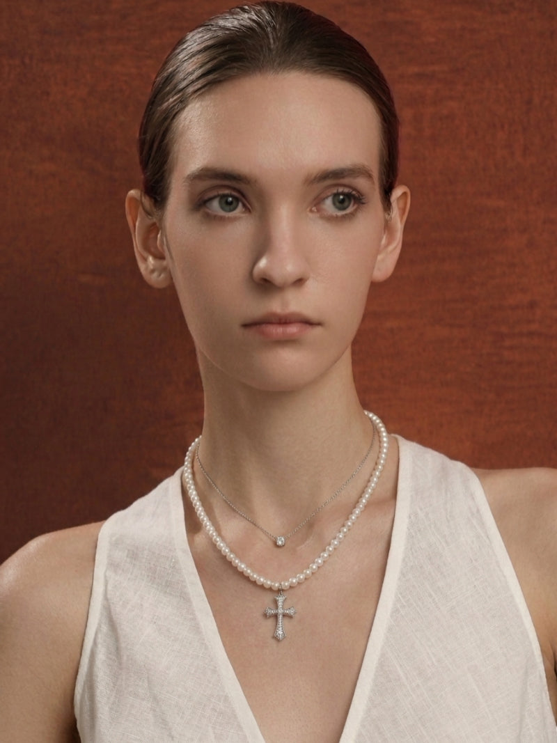 A model wears the Covenant Imitation Pearl Cross Layered Necklace, featuring a strand of white imitation pearls with a jeweled cross pendant and a delicate silver chain with a solitaire, @model