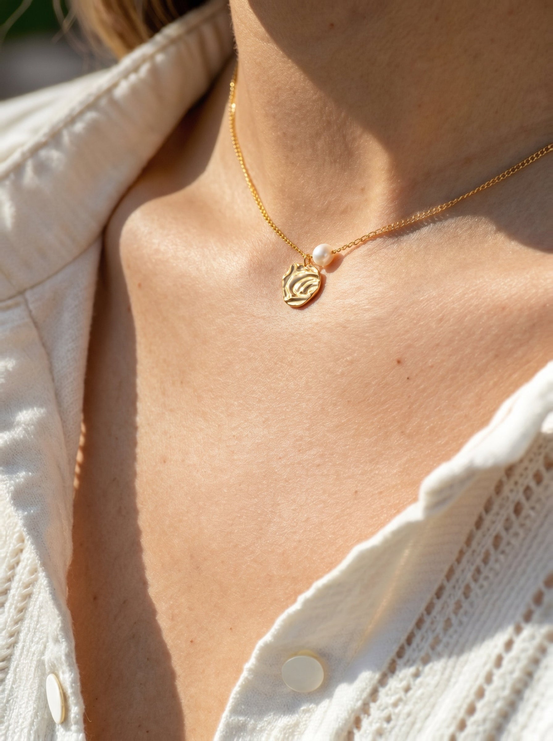 Close-up of a gold-tone Crest necklace with a single freshwater pearl and an abstract organic pendant, worn by a model with fair skin, against a white textured shirt. @model