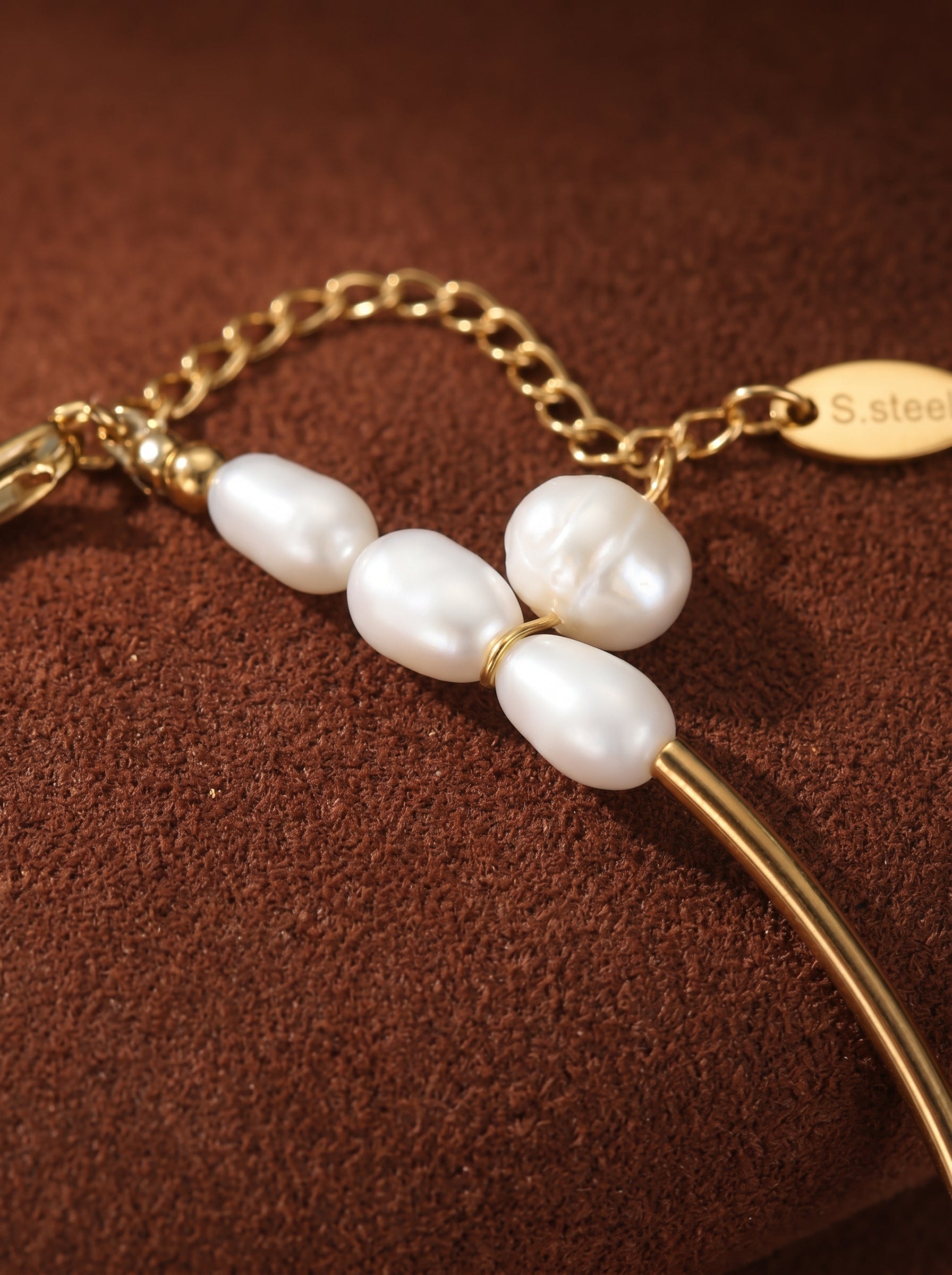 Close-up of a gold-toned chain bracelet featuring three lustrous, seed-shaped freshwater pearls and a small oval tag engraved with 'S.steel' on a textured brown surface.@pearl