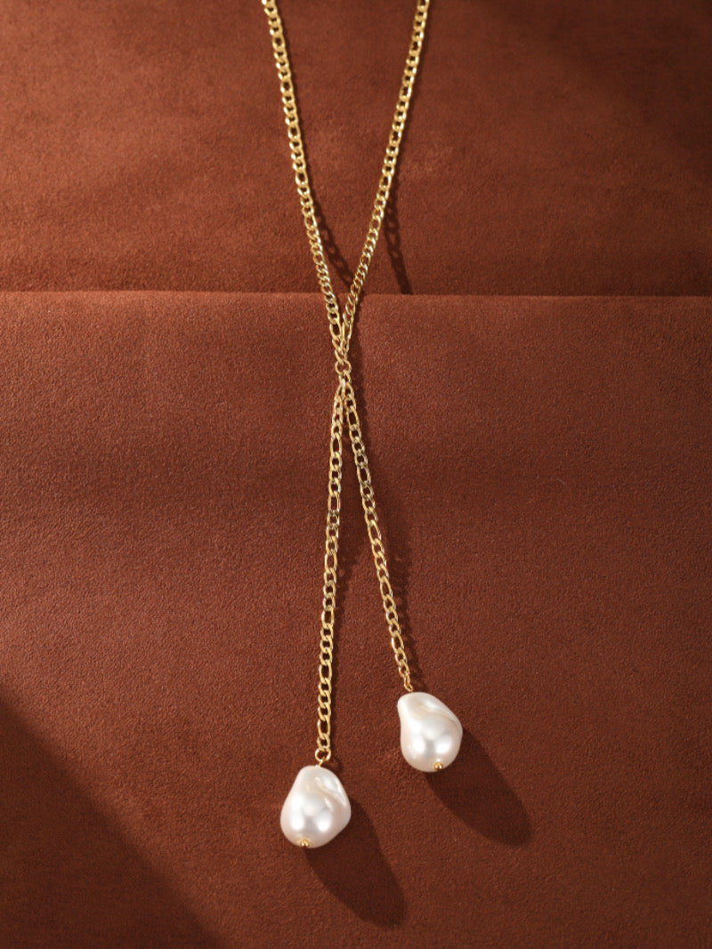 Gold-toned chain necklace with a lariat design featuring two irregular white imitation Baroque pearls hanging at the ends, displayed on a brown textured background.