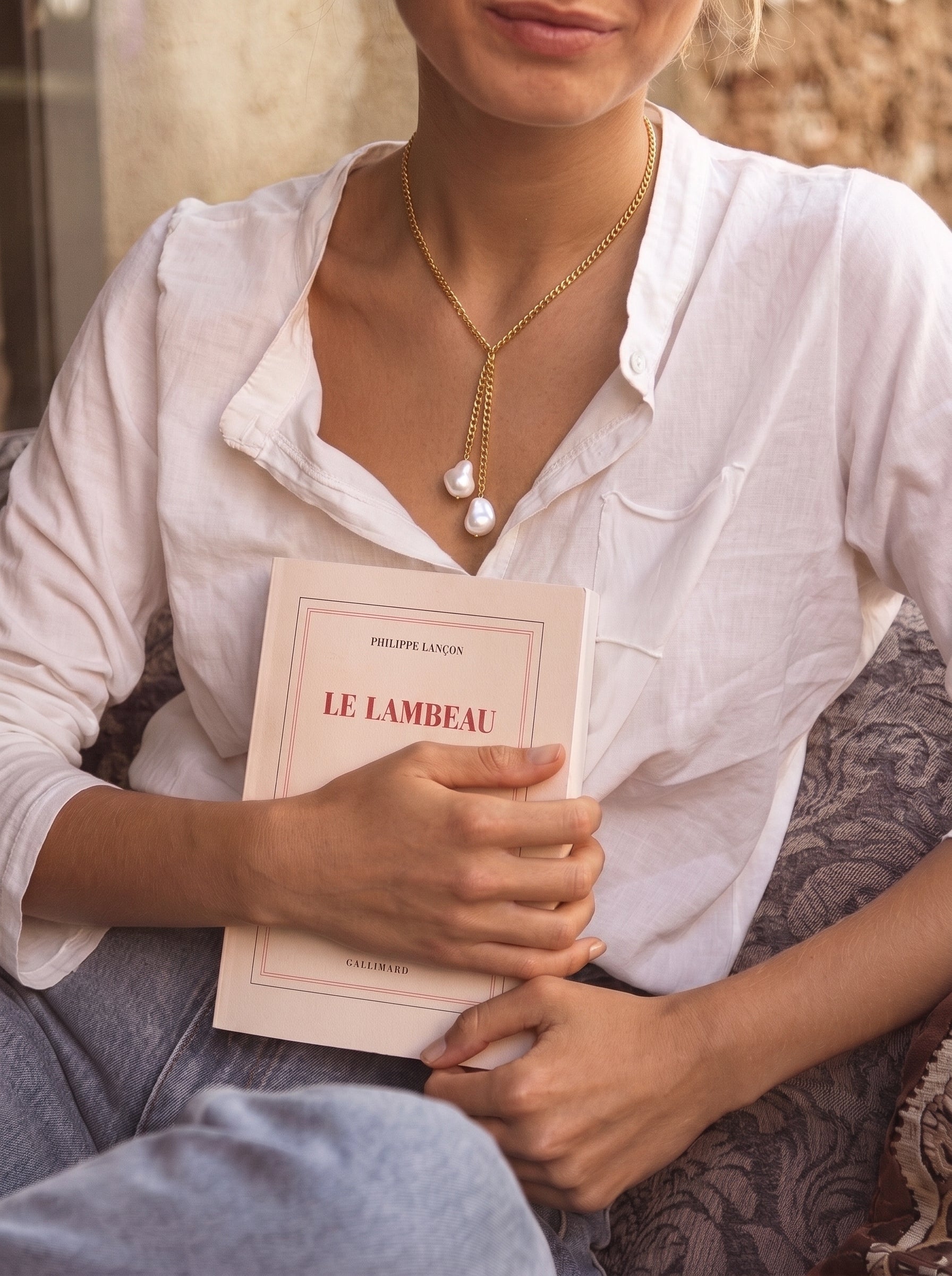 A woman in a white shirt holds a book while wearing a gold lariat necklace with two irregular white imitation pearls dangling. @lifestyle