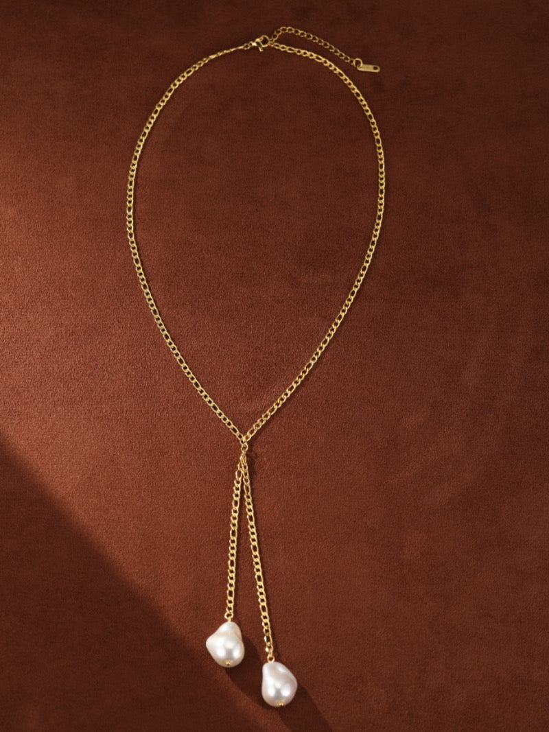 Gold-toned lariat necklace with two irregular white imitation Baroque pearls hanging from delicate chains against a brown background.