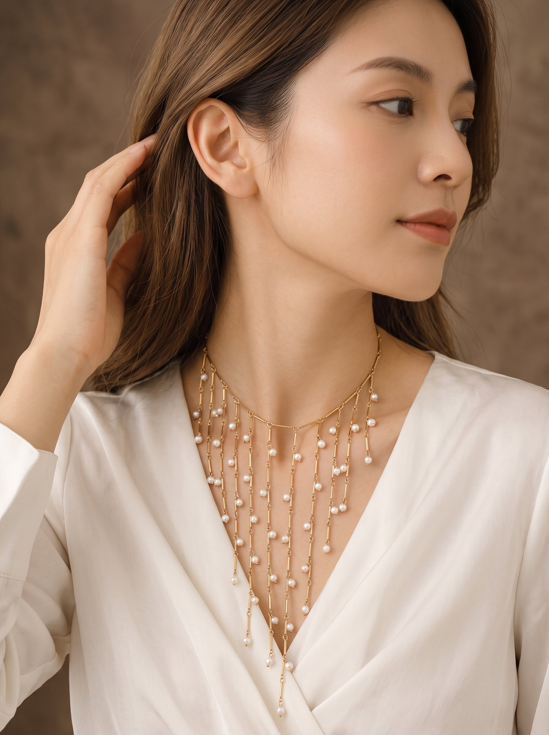 Long collier Dewfall présentant de multiples rangs dorés ornés de petites perles d'eau douce cascadant sur sa poitrine, stylisé avec un haut blanc à col en V. @lifestyle