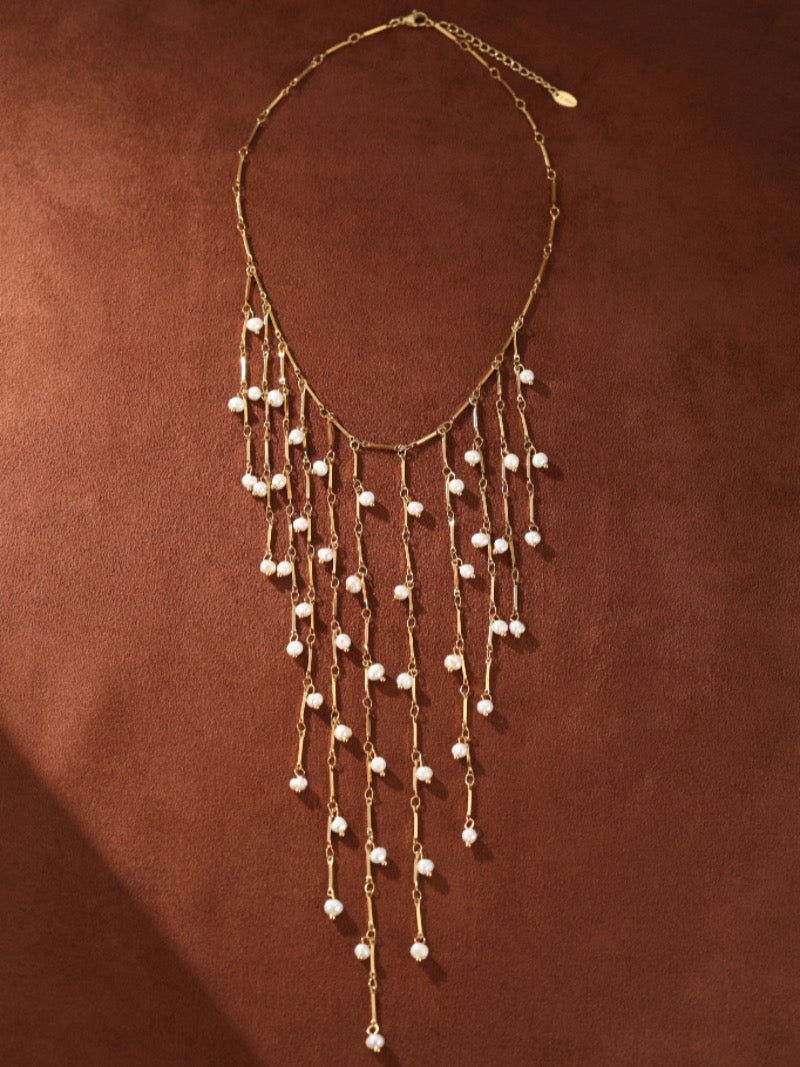 Un délicat collier Dewfall ton or présentant de multiples rangs en cascade de fines barres métalliques ornées de lumineuses perles graines d'eau douce de 3 mm, sur fond texturé marron.