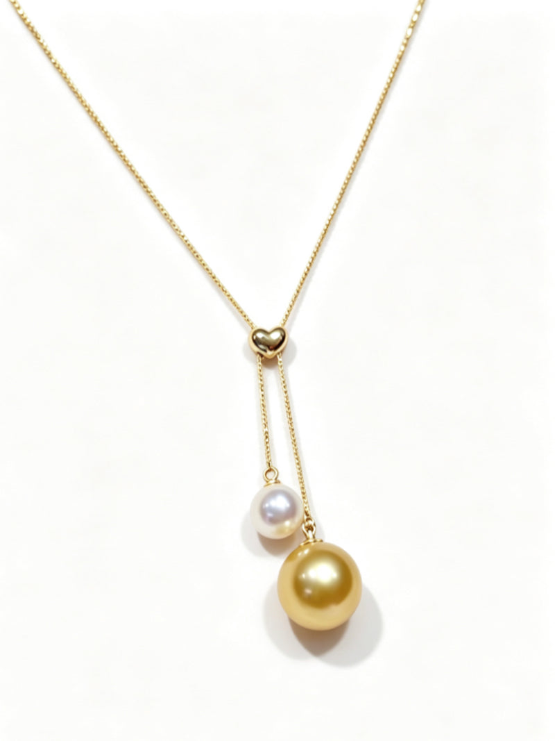 A delicate gold necklace featuring a heart-shaped clasp, with a smaller white Akoya pearl and a larger golden South Sea pearl dangling below. pearl drop pearl suspended below. @perle