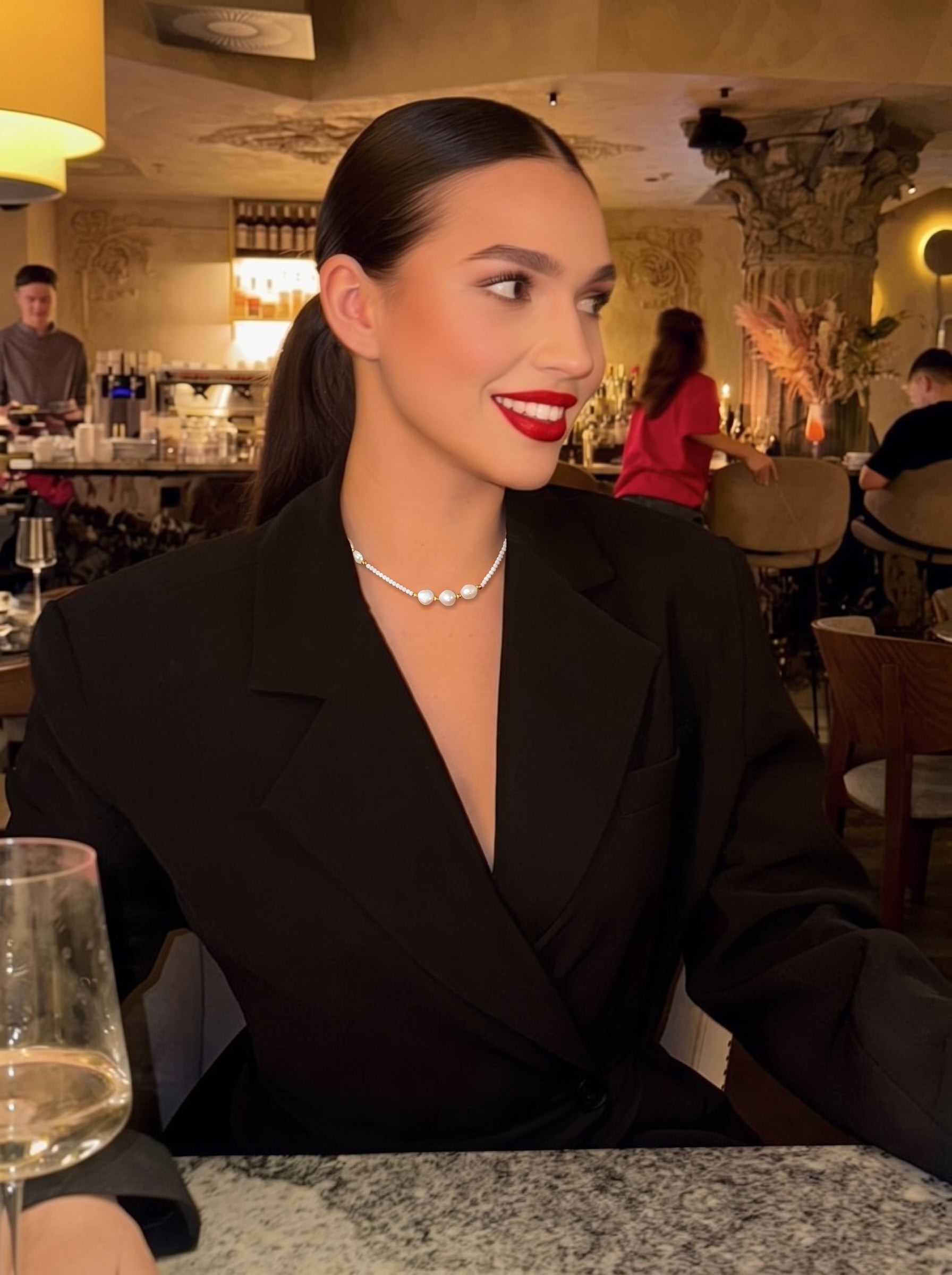 Une femme portant un blazer noir et un délicat collier à rang perlé en perle d'eau douce avec des accents dorés, assise à une table dans un restaurant. @lifestyle