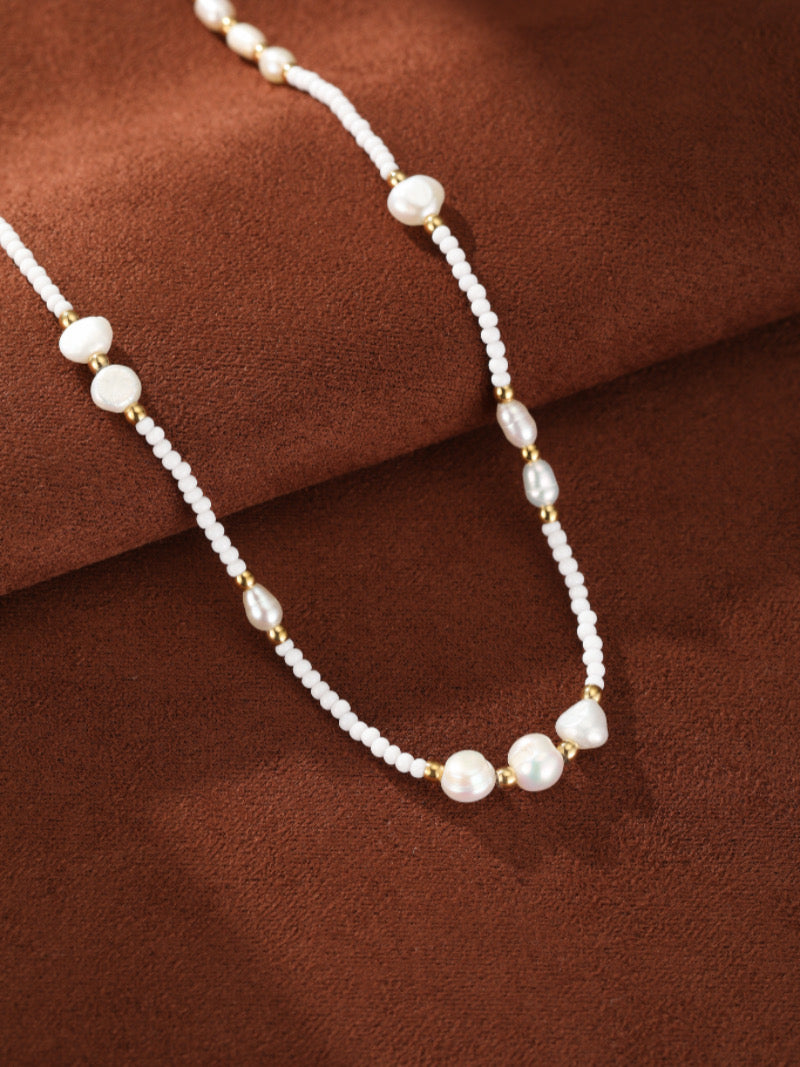 Collier Dune présentant un rang de petites perles graines blanches entrecoupées de perles baroques irrégulières d'eau douce et d'accents ton or sur fond texturé marron.