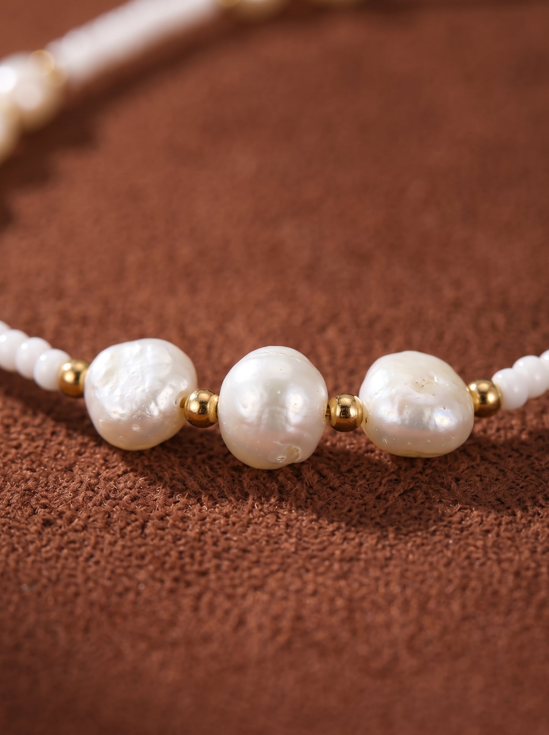 Gros plan d'un collier à rang perlé Dune en perle d'eau douce présentant des perles baroques irrégulières entrecoupées de minuscules perles graines blanches et d'accents ton or sur fond texturé marron. @pearl