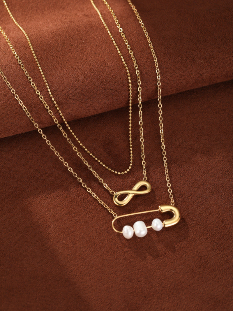 Collier superposé ton or présentant un délicat symbole infini et une breloque épingle de sûreté tenant trois lustrées perles blanches d'eau douce sur fond marron.