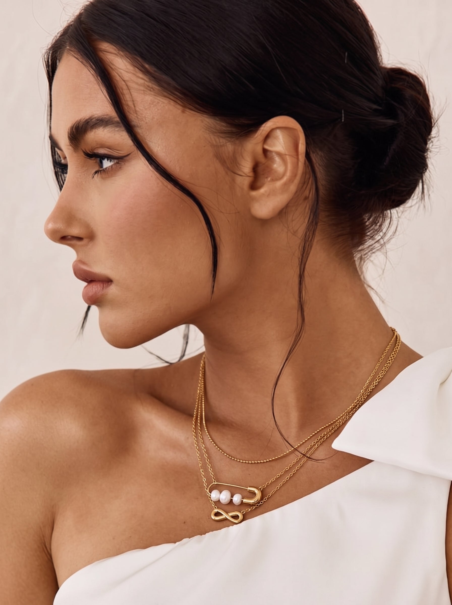 Photo de profil en gros plan d'une femme portant un délicat collier superposé doré présentant une breloque symbole infini et une breloque épingle de sûreté retenant trois lustrées perles blanches d'eau douce. @model
