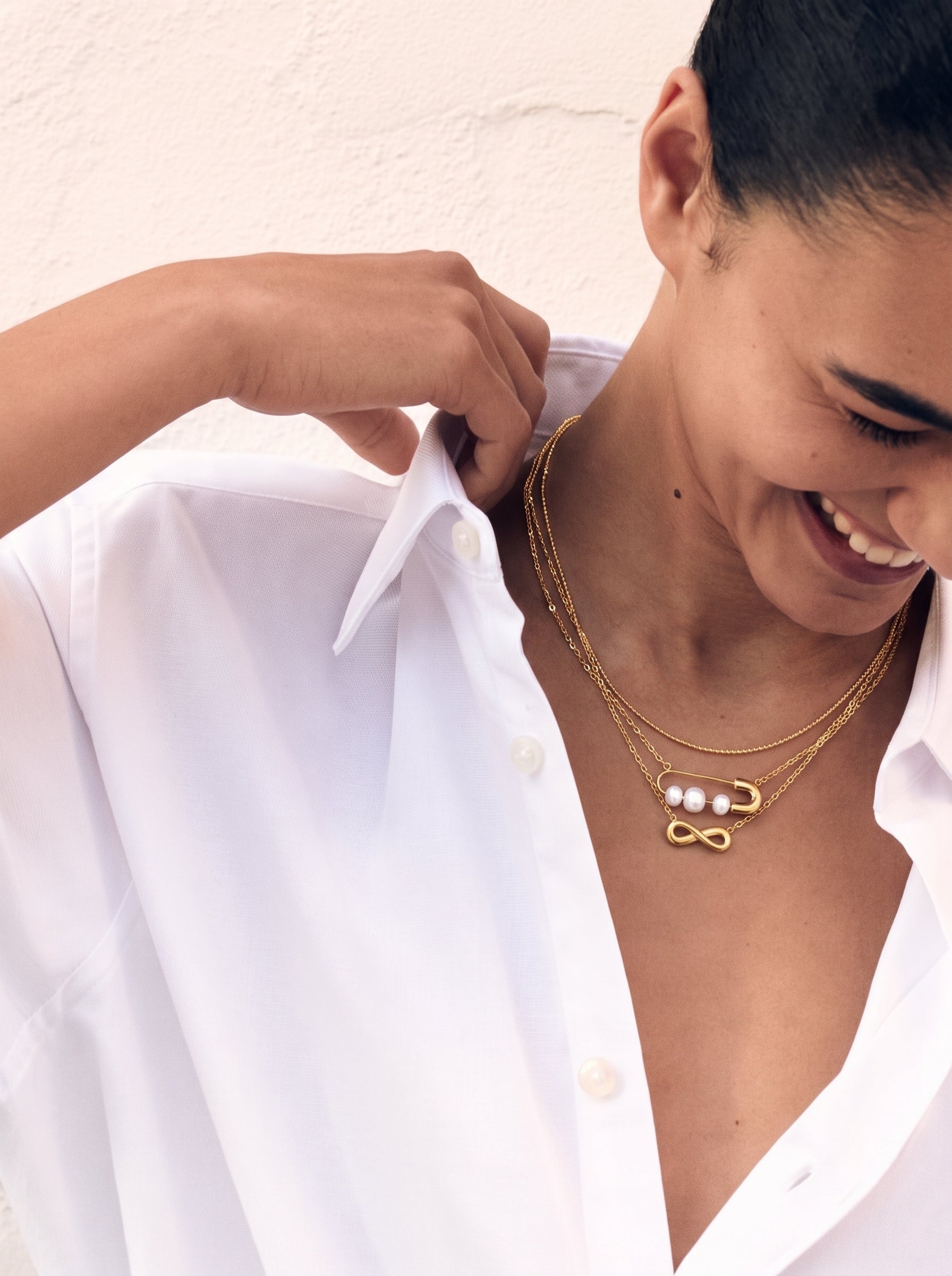 Une femme souriante porte une chemise blanche boutonnée et le collier superposé infini Endless en perle d'eau douce, présentant un symbole infini doré et une breloque épingle de sûreté avec trois perles blanches. @lifestyle