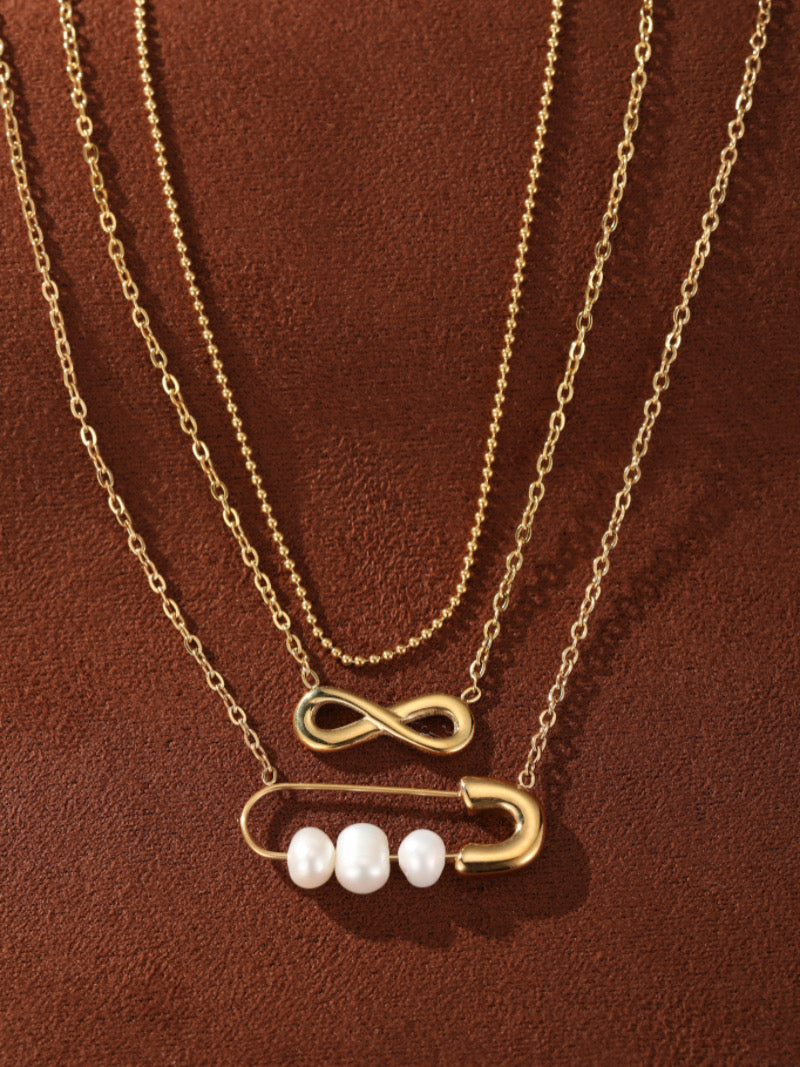 Collier superposé doré avec un symbole infini sur une chaîne et une breloque épingle de sûreté tenant trois lustrées perles graines blanches d'eau douce sur une autre chaîne, présenté sur fond texturé marron.