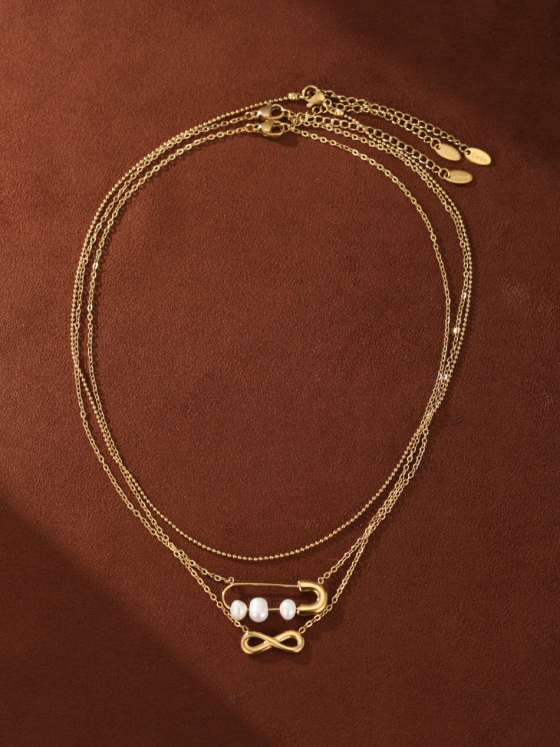 Collier superposé doré présentant une délicate chaîne avec un symbole infini et une autre chaîne avec une breloque épingle de sûreté tenant trois perles graines blanches d'eau douce.