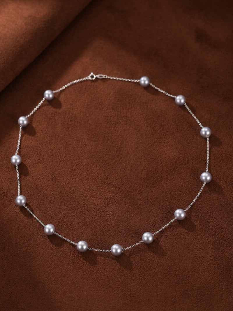 A delicate silver chain necklace featuring evenly spaced lustrous grey Akoya pearls against a textured brown background.