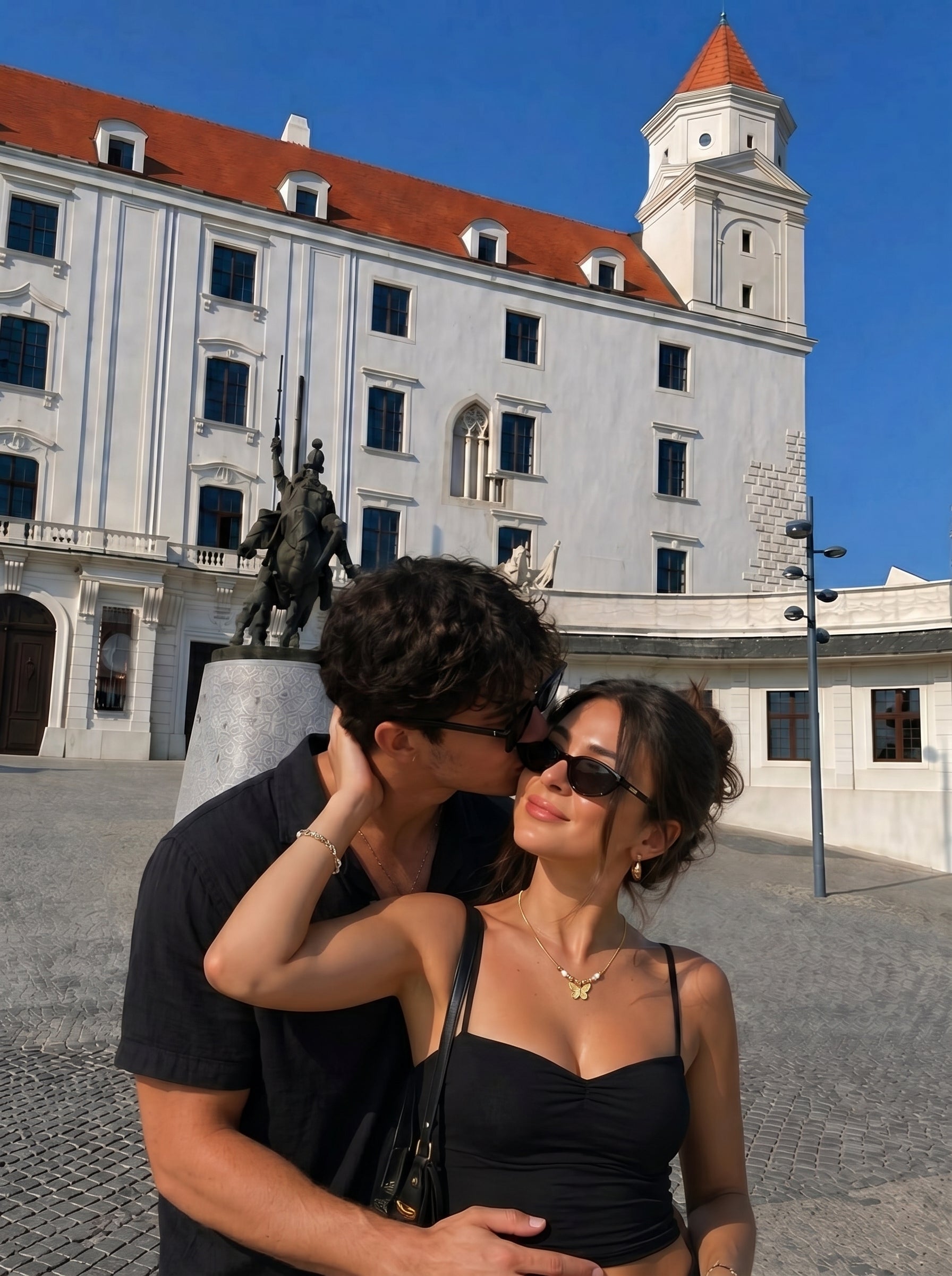 Une femme sourit tandis qu'un homme l'embrasse sur la joue, révélant le collier pendentif unique papillon Ephemeral Dance en perle d'eau douce avec son délicat papillon doré et ses détails en perle. Le couple se tient devant un bâtiment blanc au toit de tuiles rouges. @lifestyle